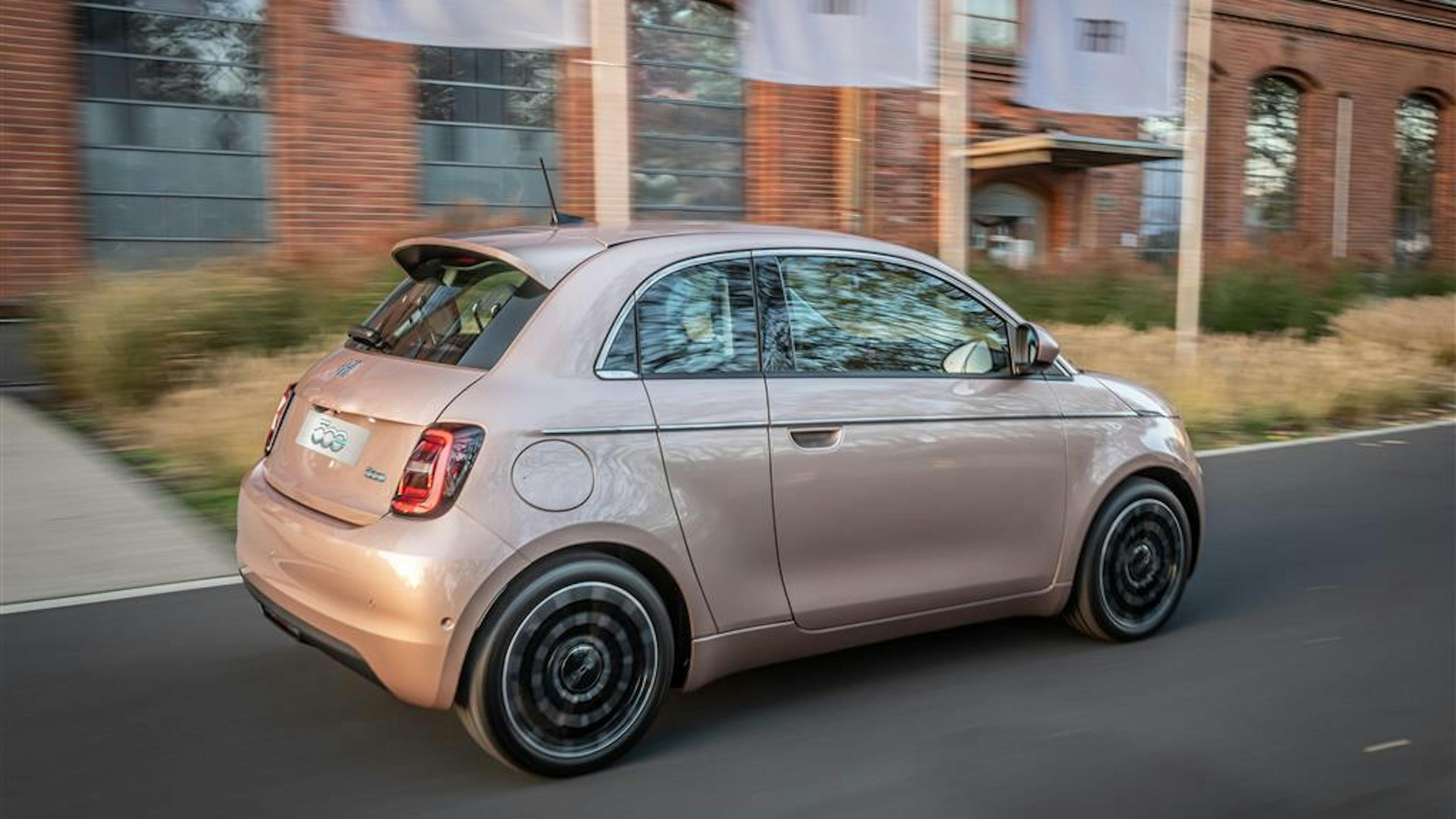 A rose-colored Fiat 500 drives from left to right in front of an industrial façade.