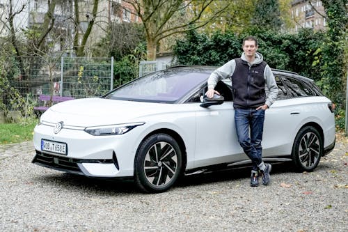 Ein Auto-Redakteur lehnt lächelnd an einem weißen VW ID.7 Tourer, geparkt auf einem Kiesplatz.