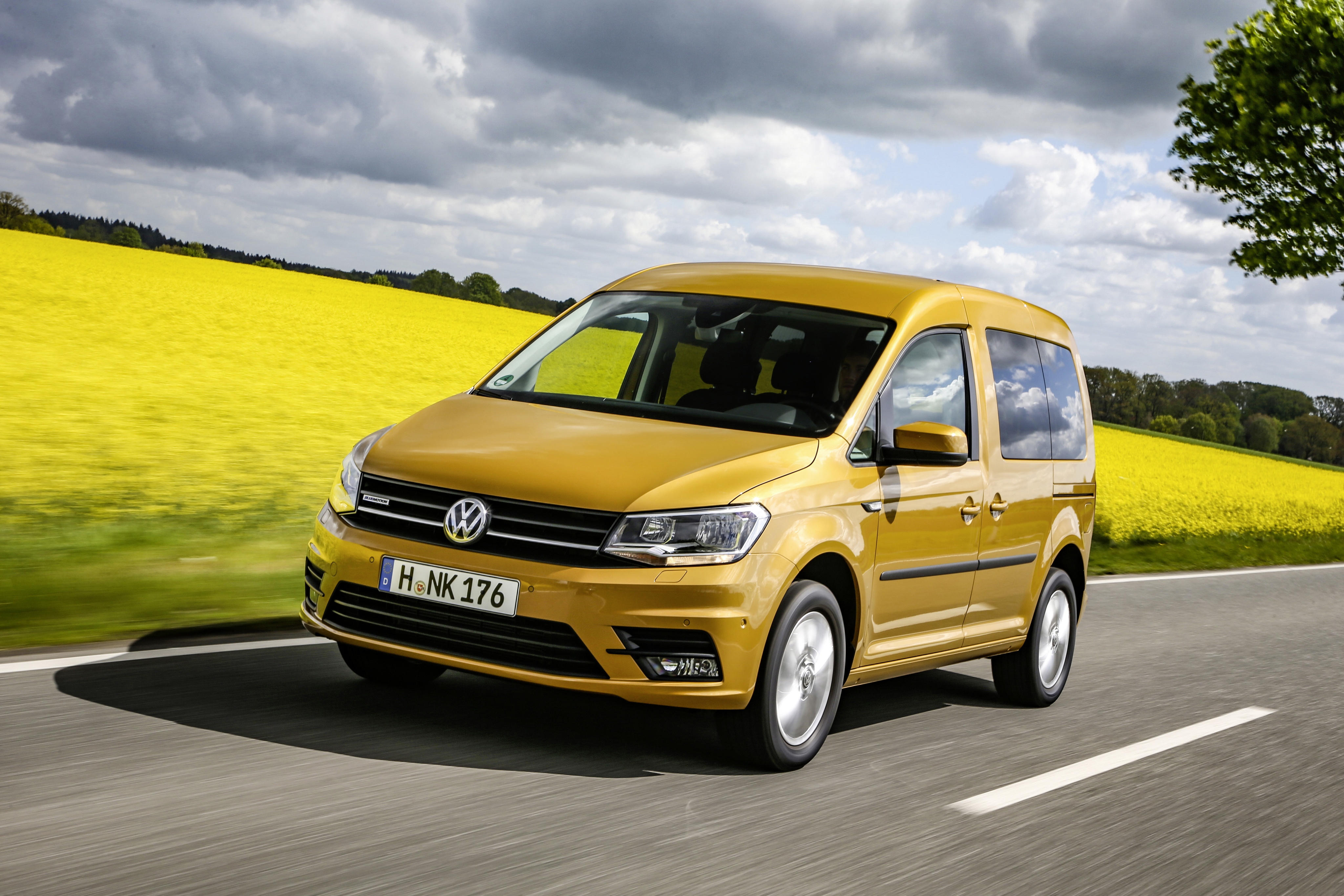 Ein bronzefarbener VW Caddy fährt auf einer Straße mit Rapsfeldern im Hintergrund.