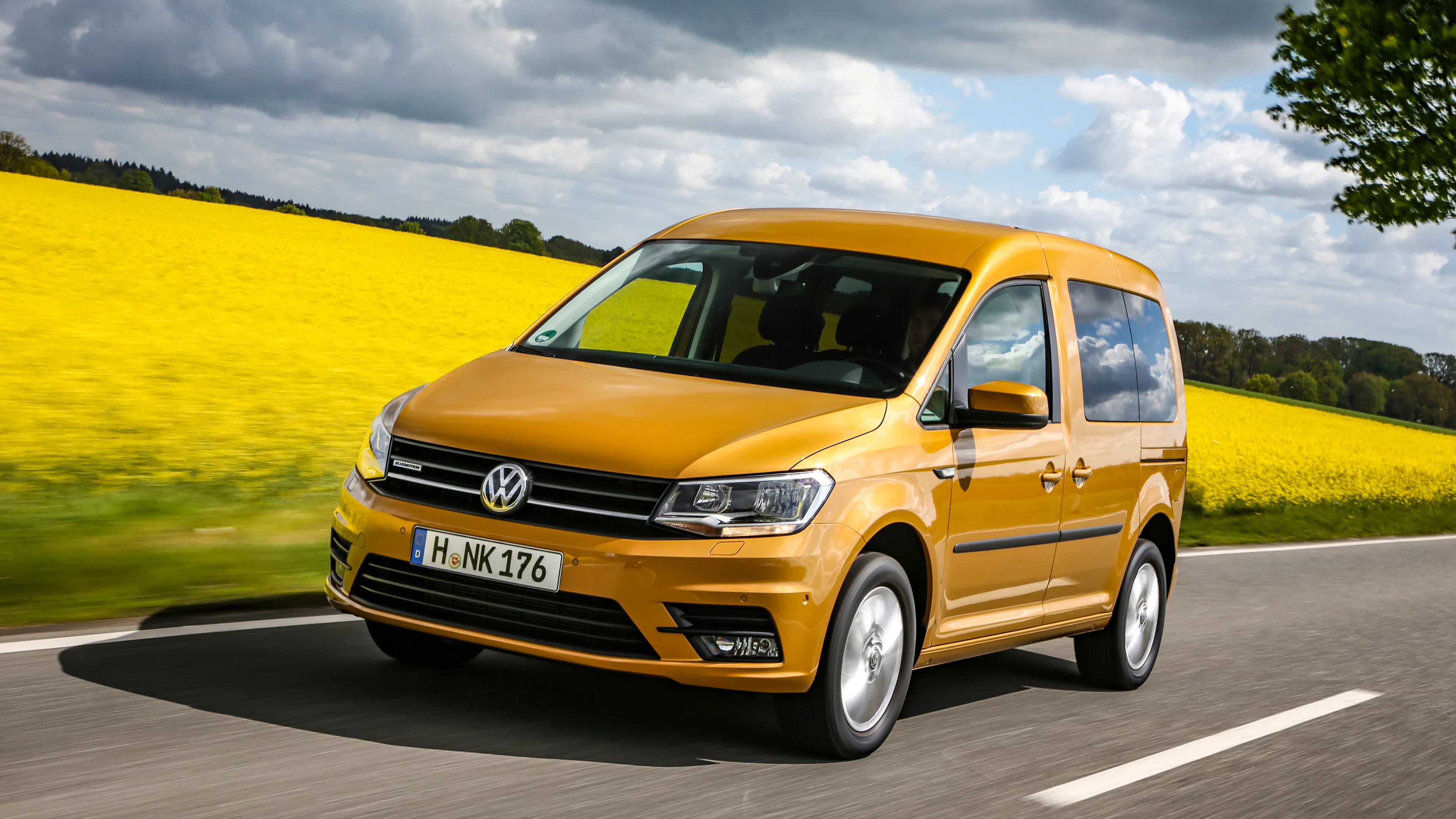 Ein bronzefarbener VW Caddy fährt auf einer Straße mit Rapsfeldern im Hintergrund.