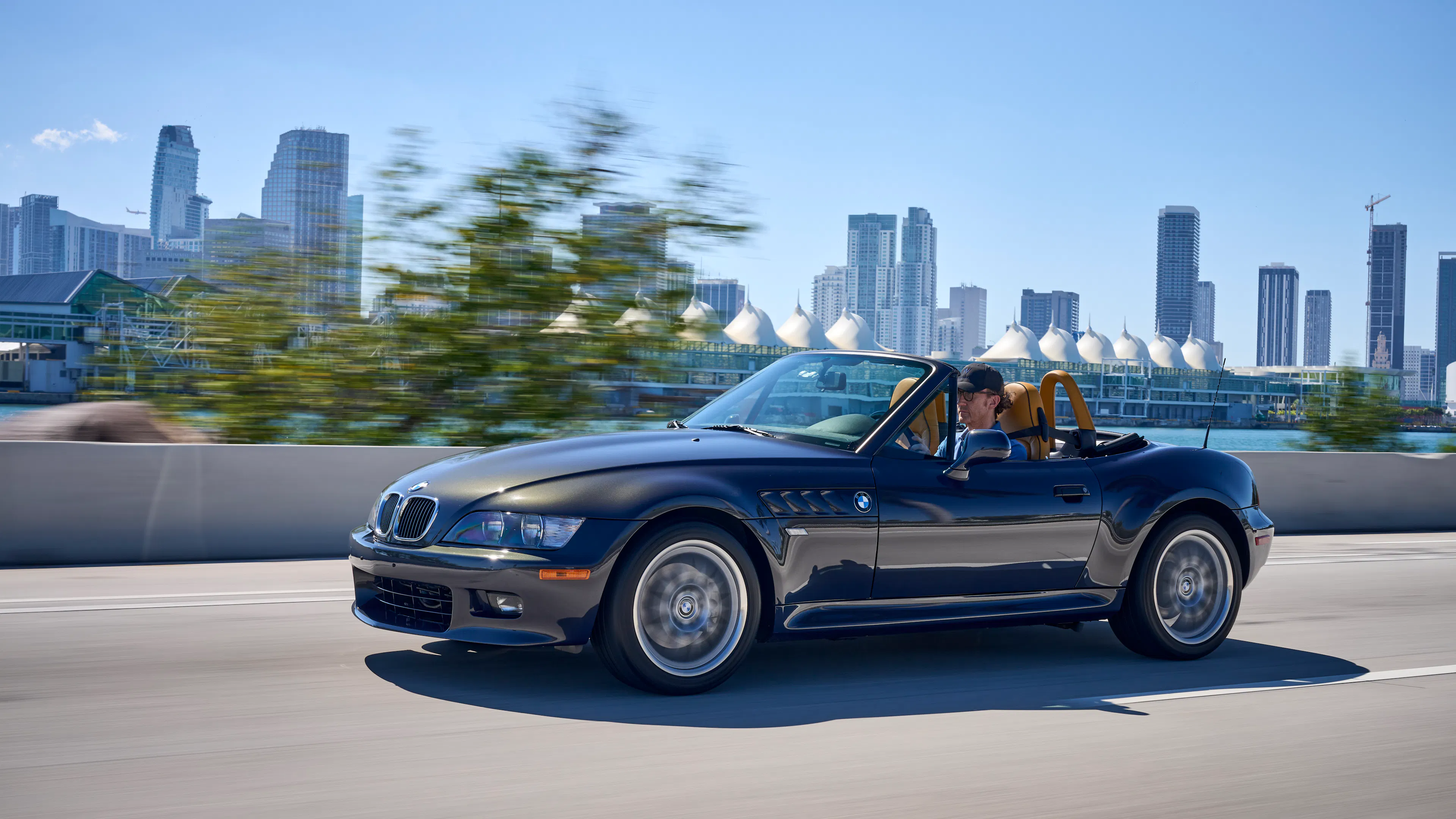 Ein offener BMW Z3 fährt vor einer Hochhaus-Skyline entlang.