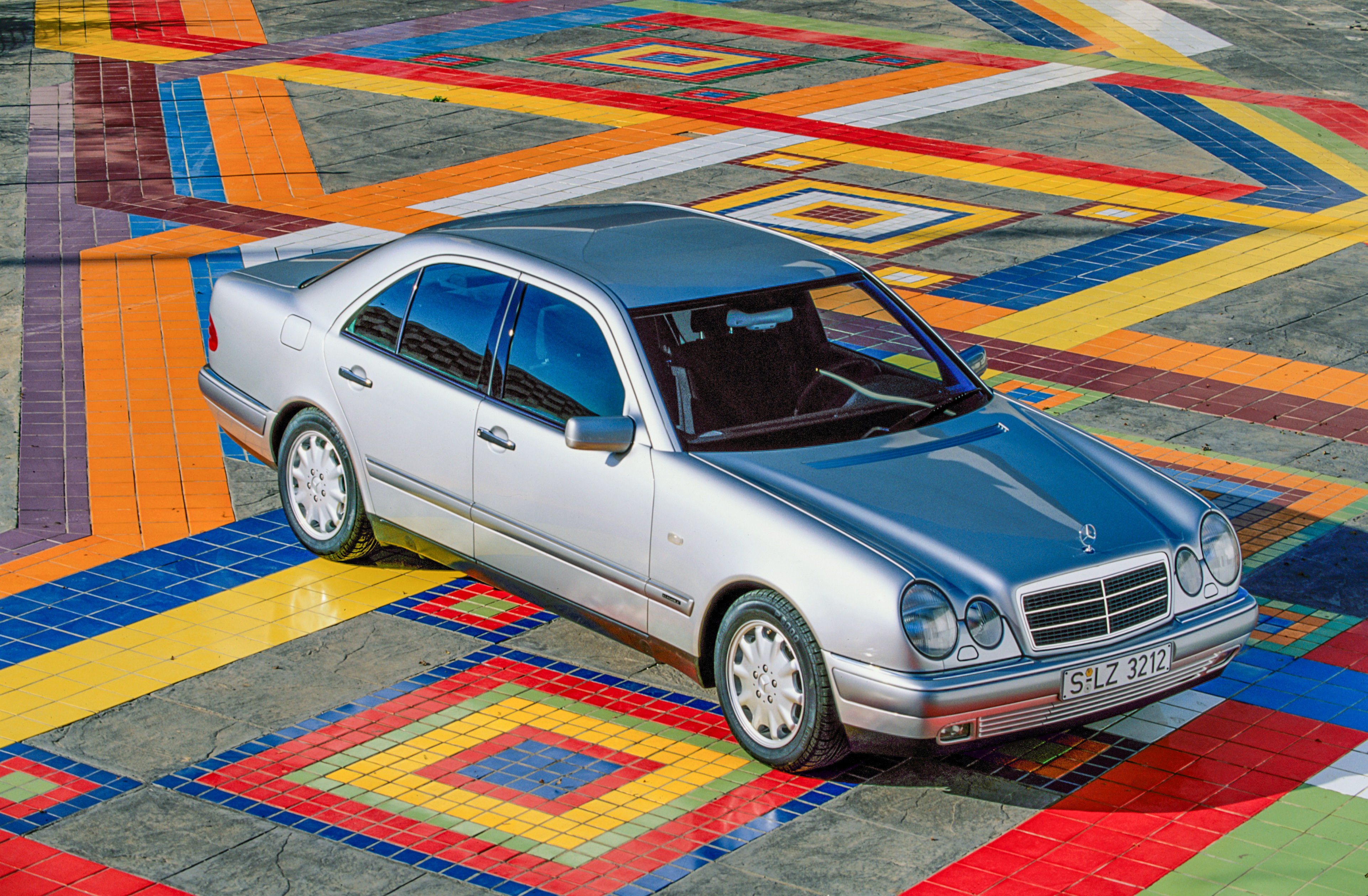 Ein silberner Mercedes der Baureihe W210 steht auf einem bunt gefliesten Platz.