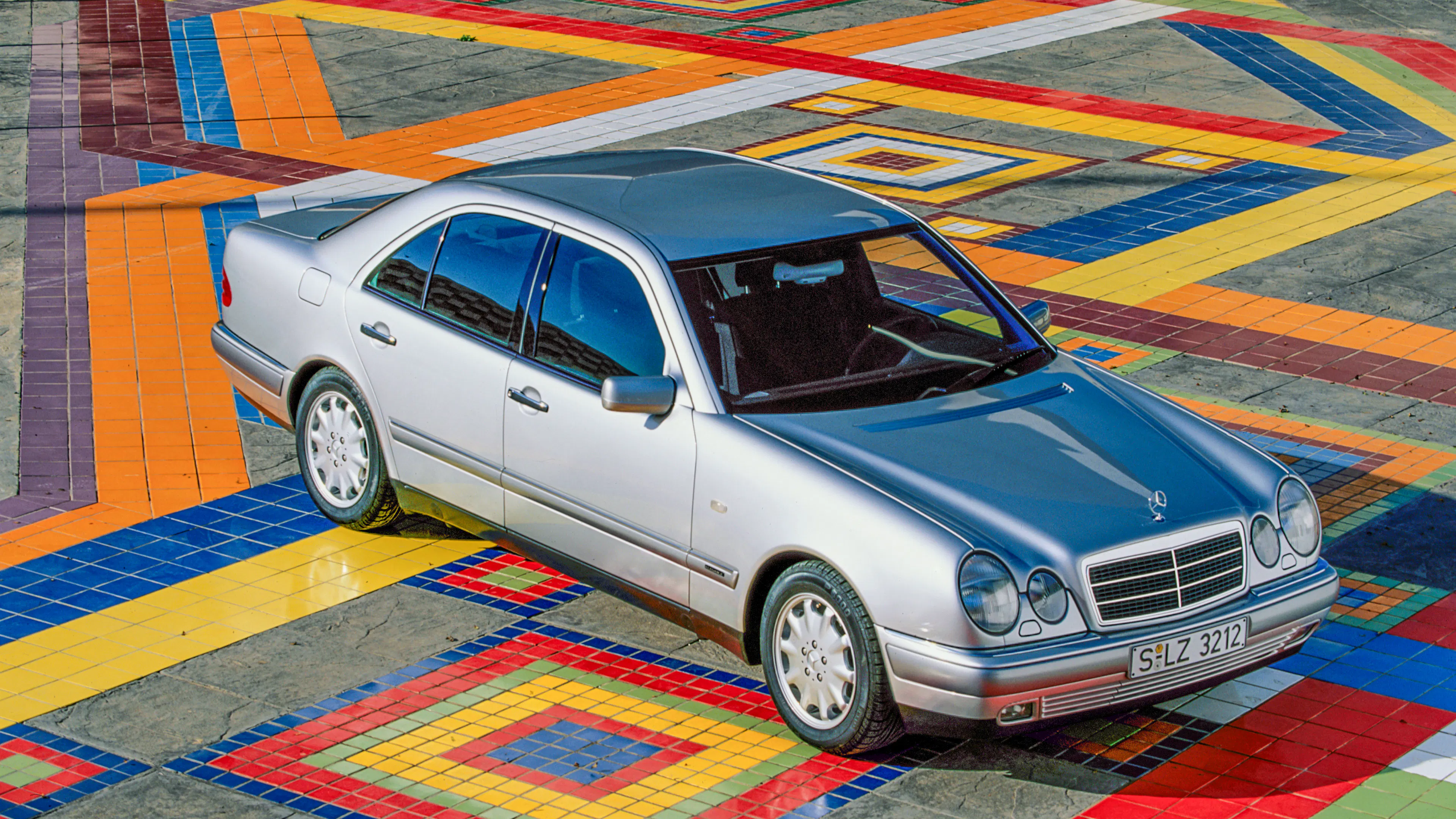 Ein silberner Mercedes der Baureihe W210 steht auf einem bunt gefliesten Platz.
