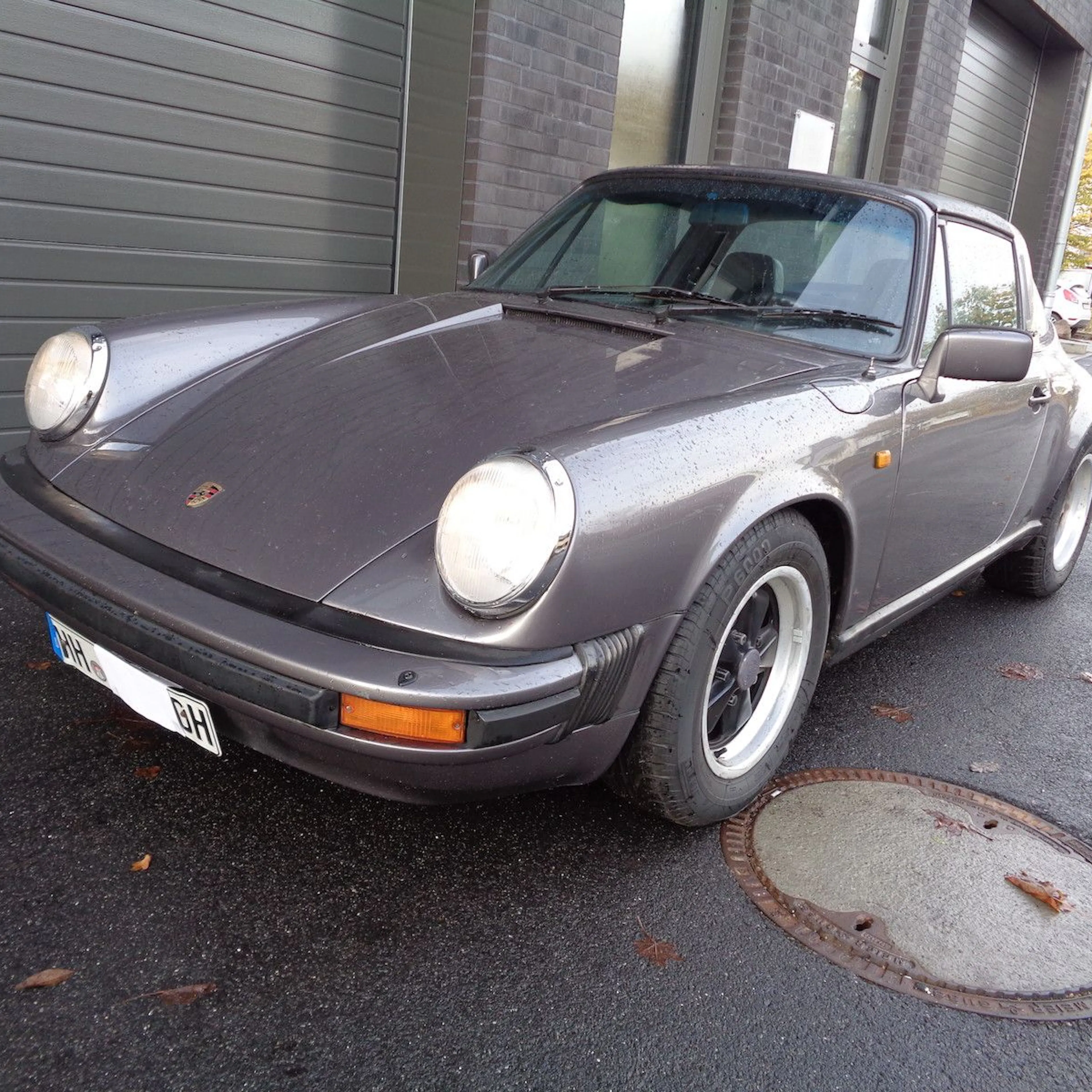 Grauer, klassischer Porsche 911 auf einem Parkplatz vor einem grauen Gebäude.