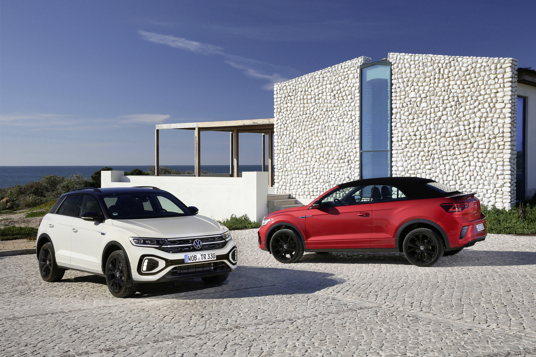 Ein weißer VW T-Roc und ein rotes VW T-Roc Cabrio stehen auf einer gepflasterten Fläche vor einem mediterranen Gebäude.