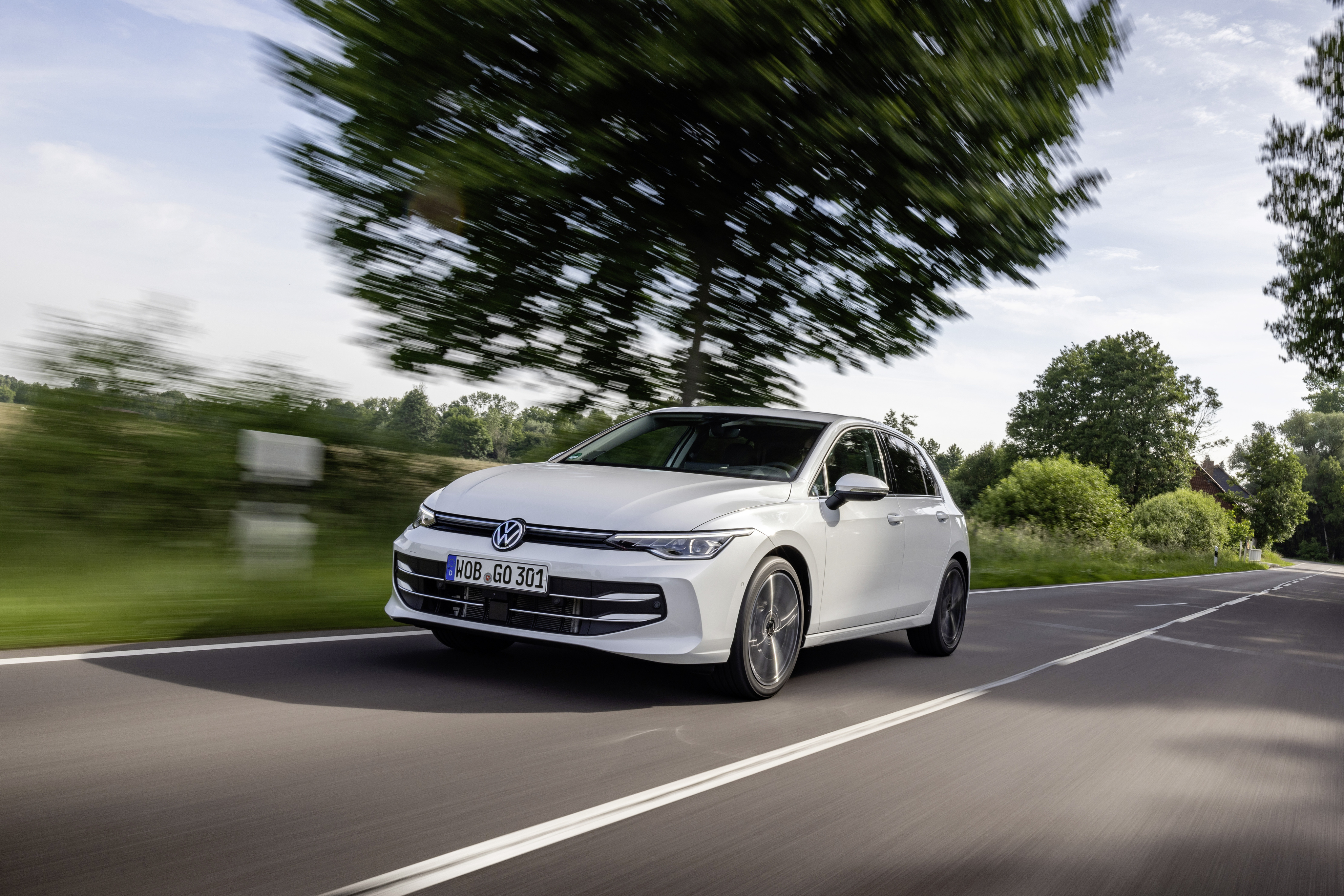 Ein weißer VW Golf fährt auf einer Landstraße mit Bäumen.