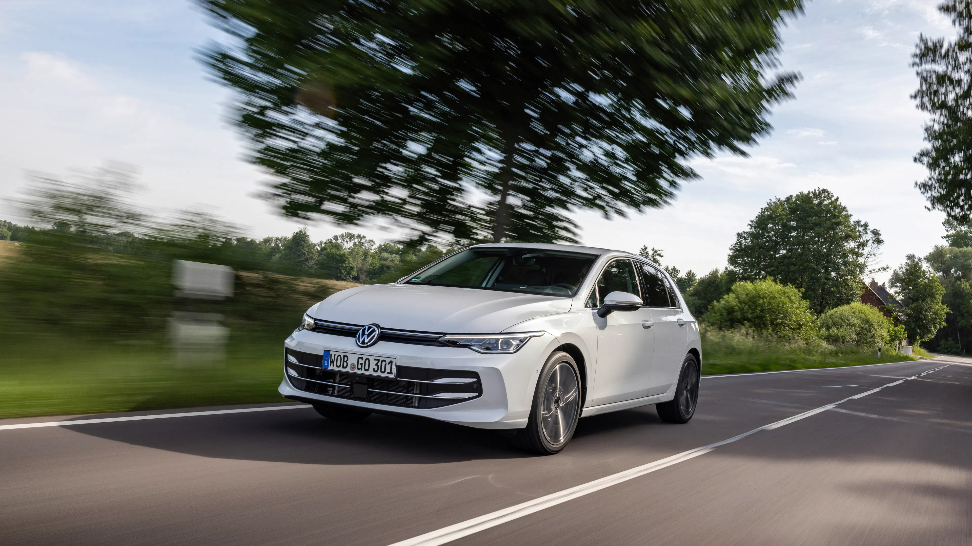 Ein weißer VW Golf fährt auf einer Landstraße mit Bäumen.