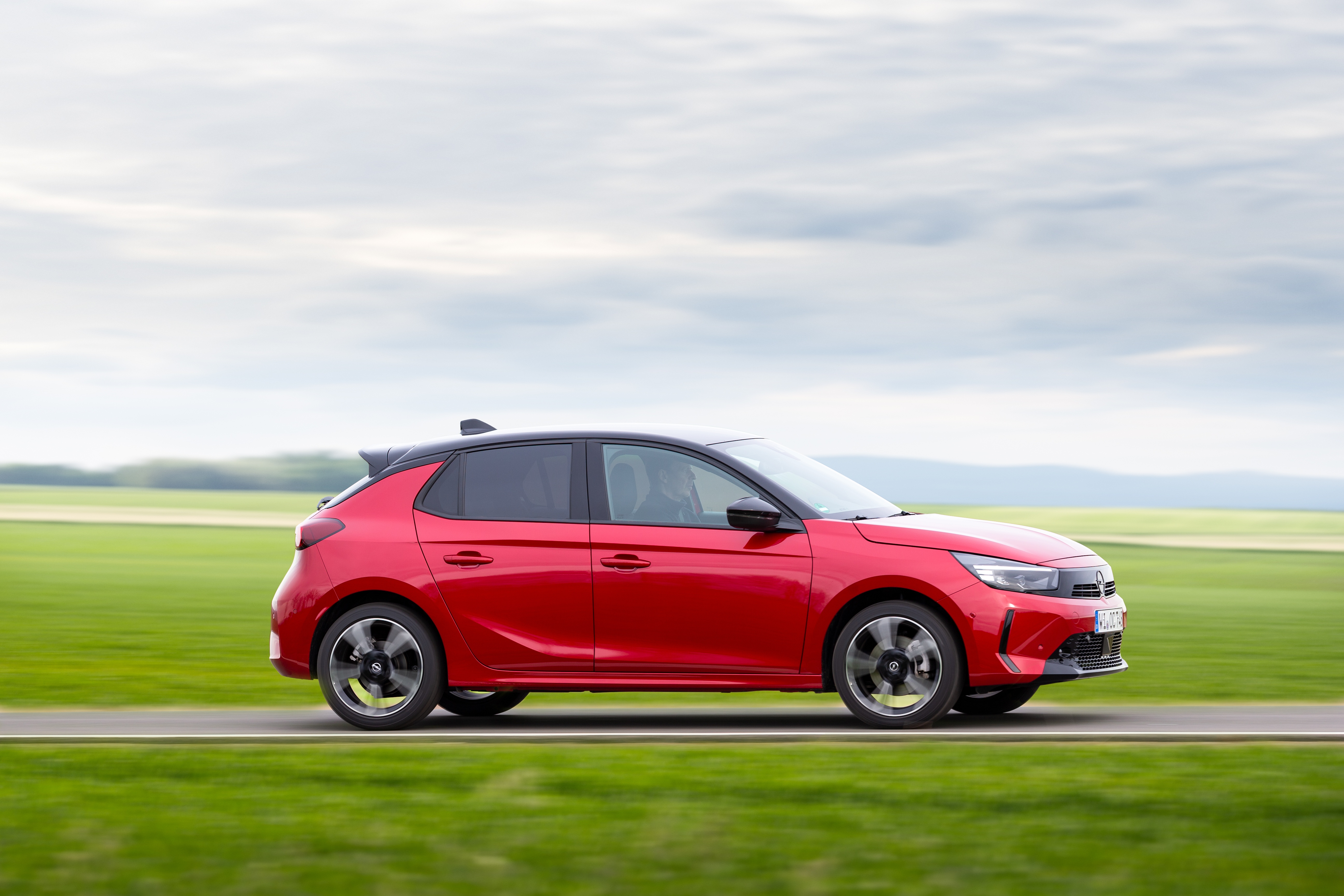 Ein roter Opel Corsa fährt auf einer Straße mit Wiesen im Hintergrund.