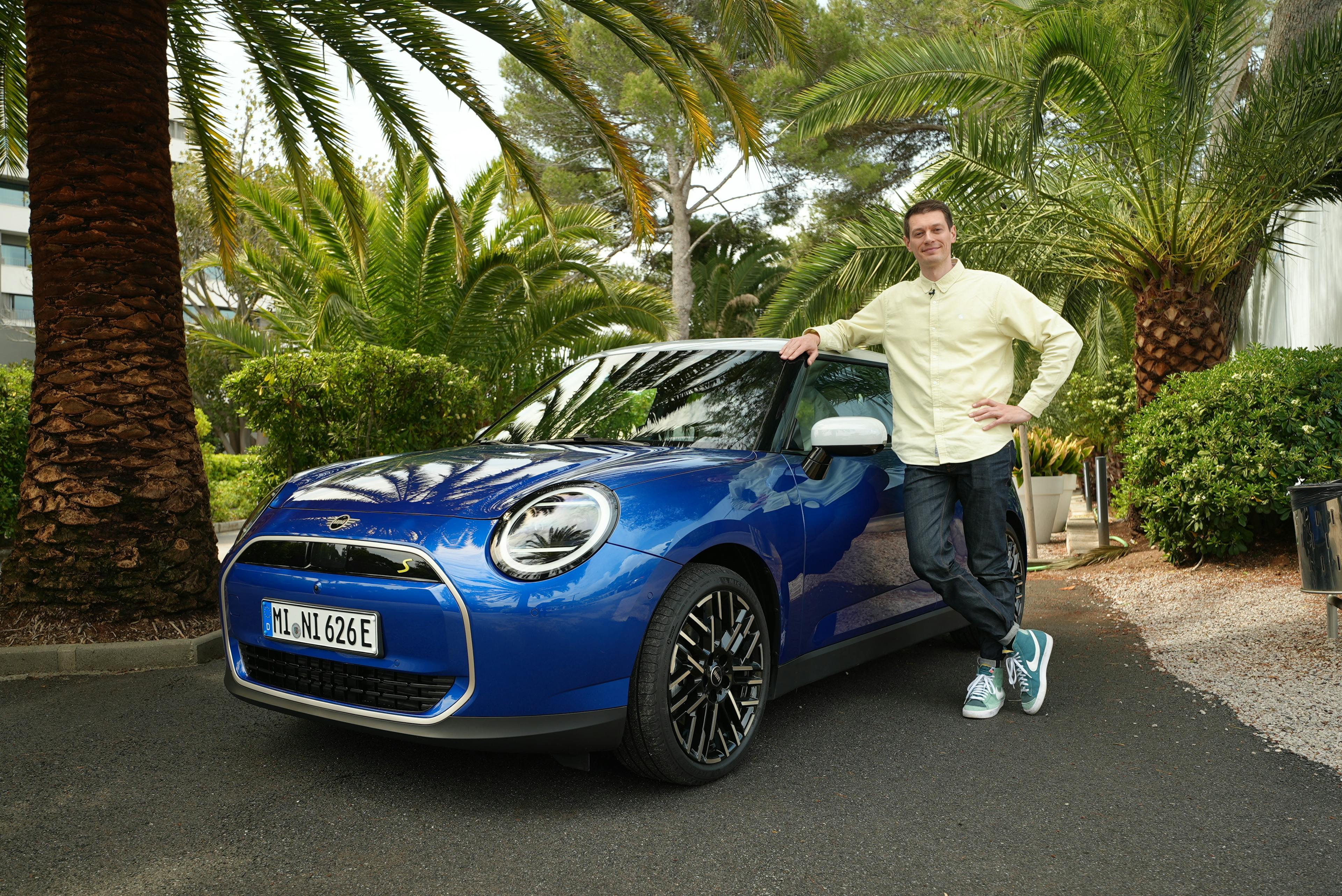 Der Autojournalist Peter R. Fischer steht neben einem blauen Mini Cooper SE vor Palmen.