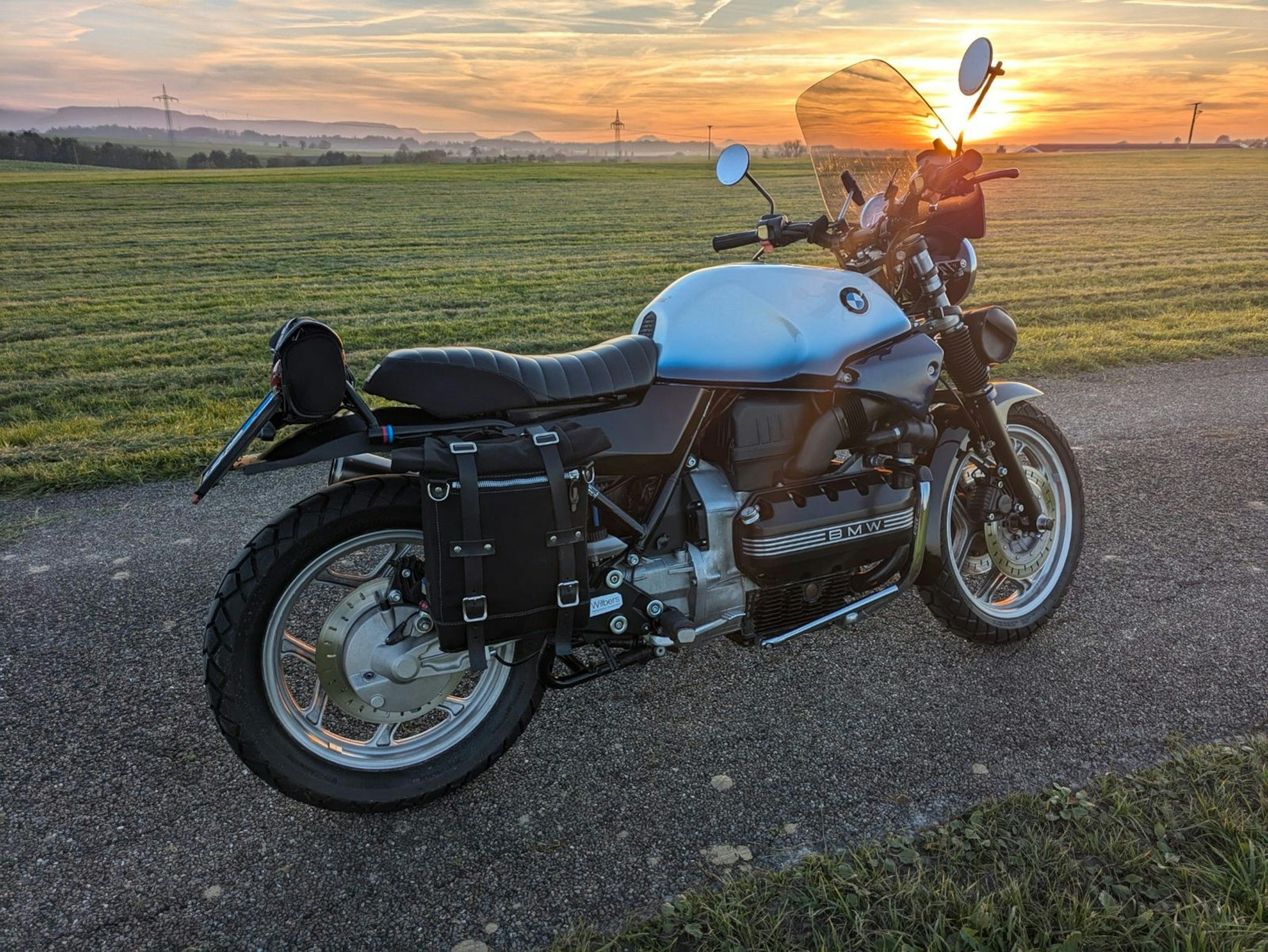 Ein Motorrad der Marke BMW mit blau-weißem Tank steht auf einem asphaltierten Weg neben einer Wiese, während im Hintergrund die Sonne über einer hügeligen Landschaft untergeht.