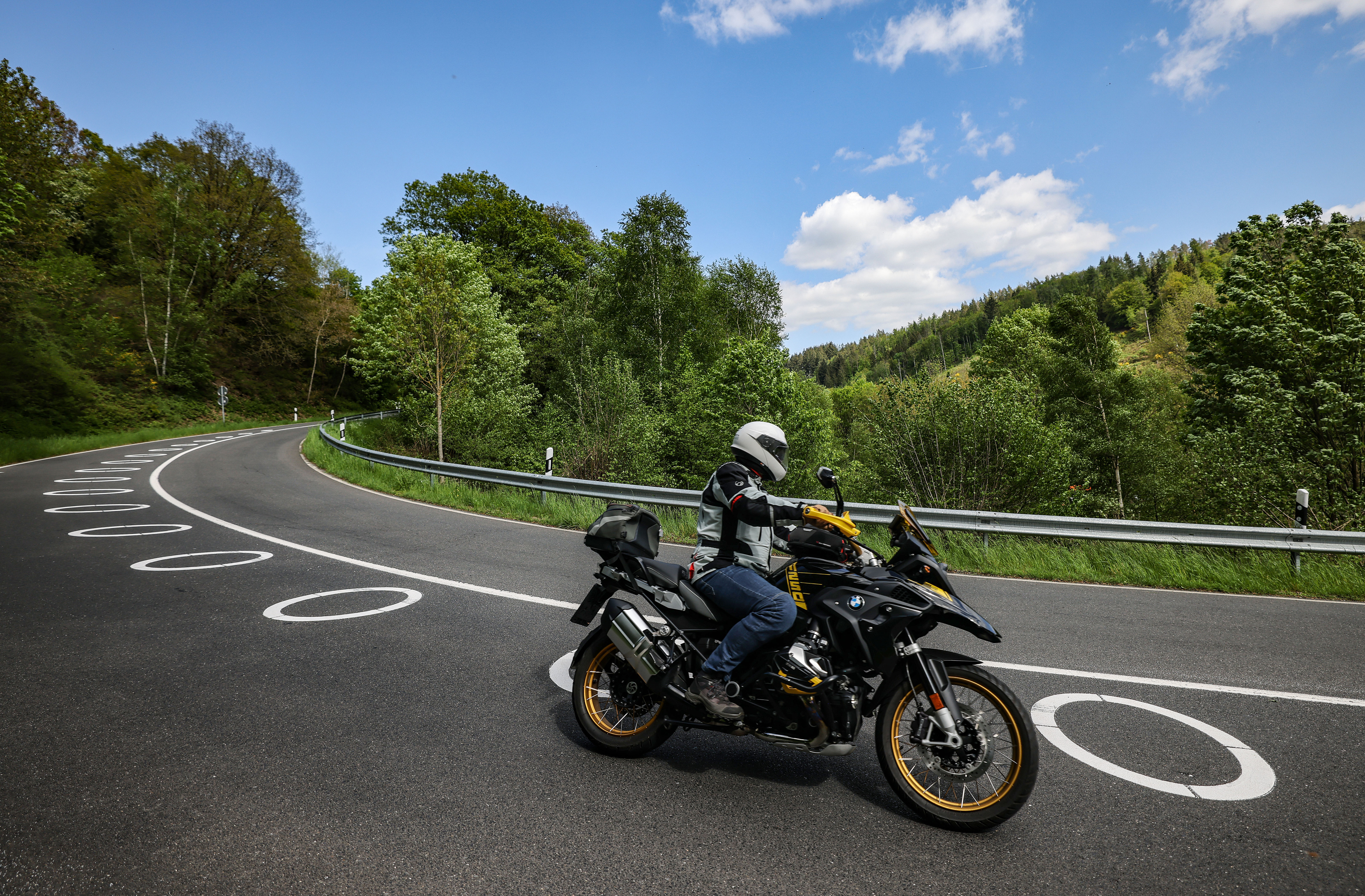 Ein Motorradfahrer mit Schutzkleidung und Helm fährt auf einer kurvigen Landstraße mit markierten Fahrbahnlinien, umgeben von grüner Landschaft.