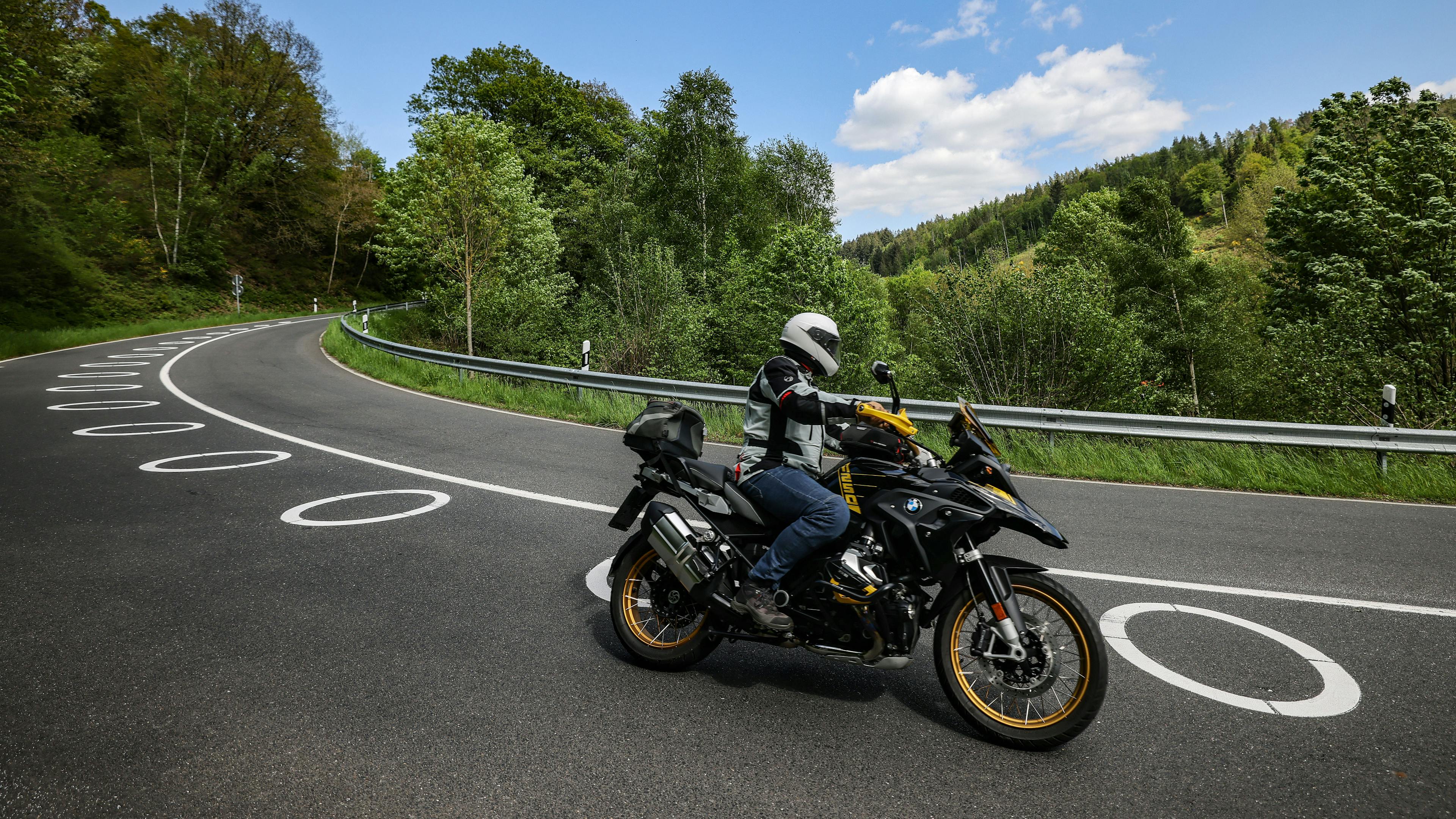 Ein Motorradfahrer mit Schutzkleidung und Helm fährt auf einer kurvigen Landstraße mit markierten Fahrbahnlinien, umgeben von grüner Landschaft.