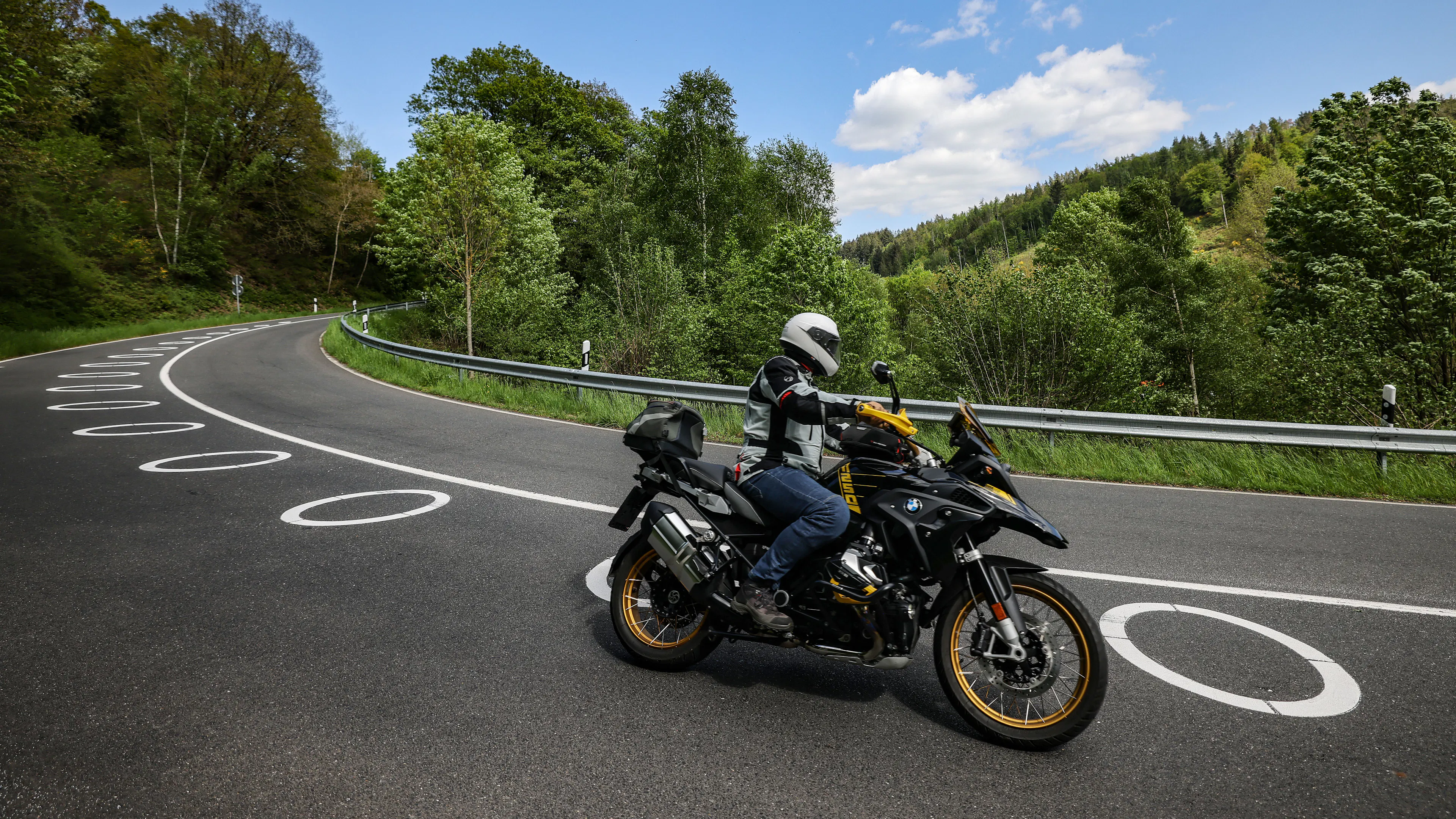 Ein Motorradfahrer mit Schutzkleidung und Helm fährt auf einer kurvigen Landstraße mit markierten Fahrbahnlinien, umgeben von grüner Landschaft.