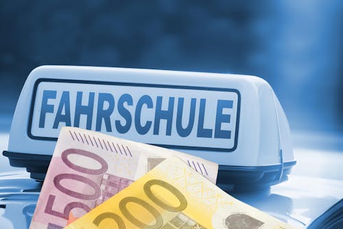  Ein Fahrschulauto mit einem „Fahrschule”-Schild auf dem Dach, im Vordergrund liegen mehrere Euro-Geldscheine.
