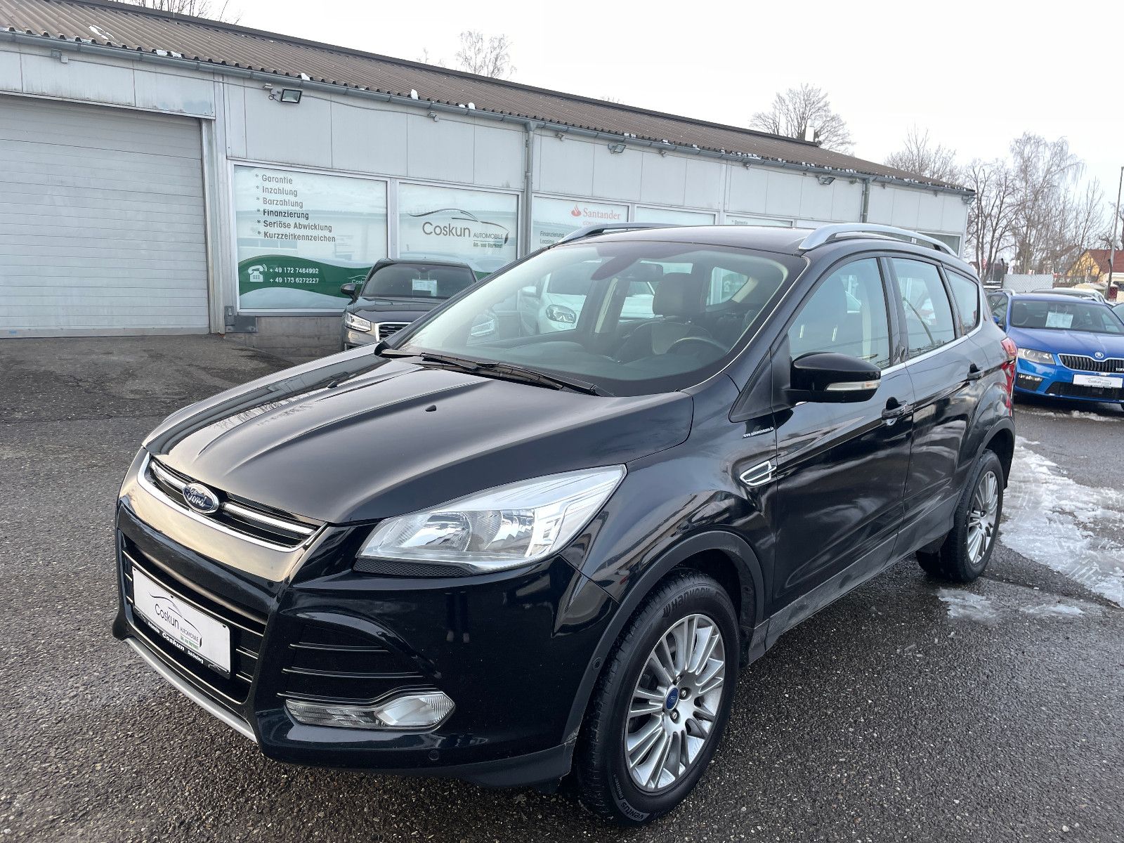 Schwarzer SUV Ford Kuga steht auf einem Parkplatz vor einer Autowerkstatt. Boden mit Schnee und Wasser bedeckt.