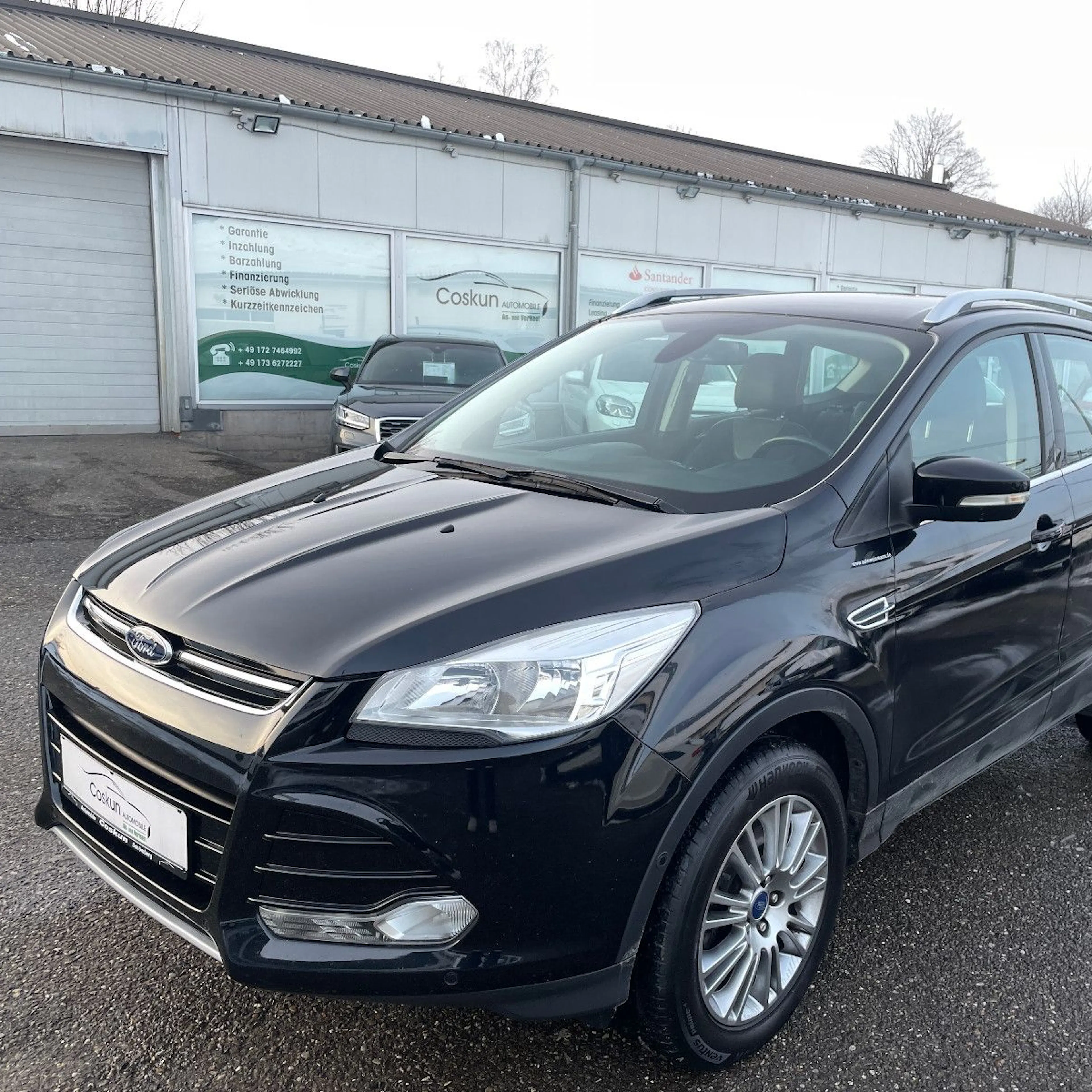 Schwarzer SUV Ford Kuga steht auf einem Parkplatz vor einer Autowerkstatt. Boden mit Schnee und Wasser bedeckt.