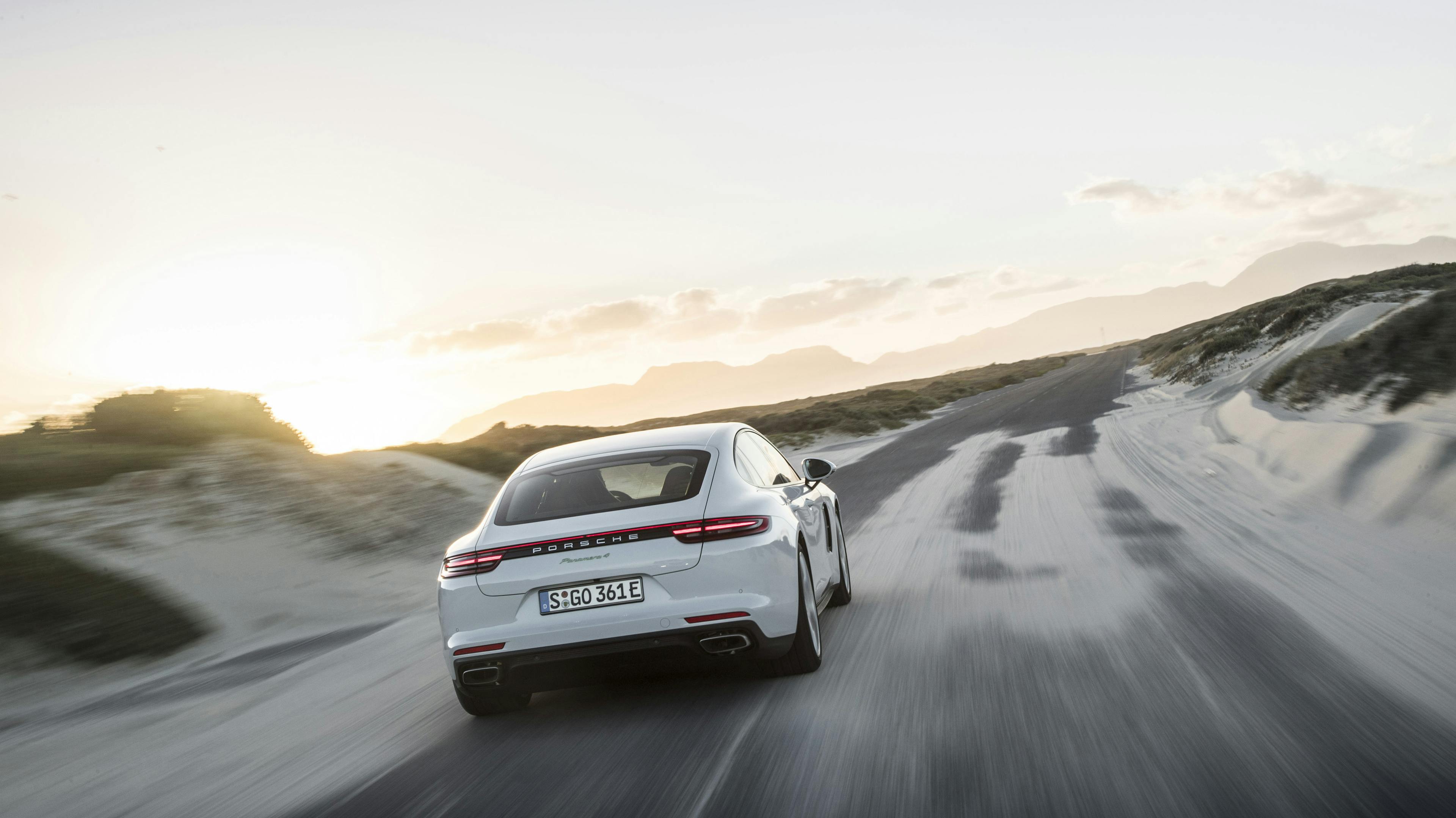  Ein weißer Porsche Panamera fährt auf einer Landstraße durch Sanddünen.