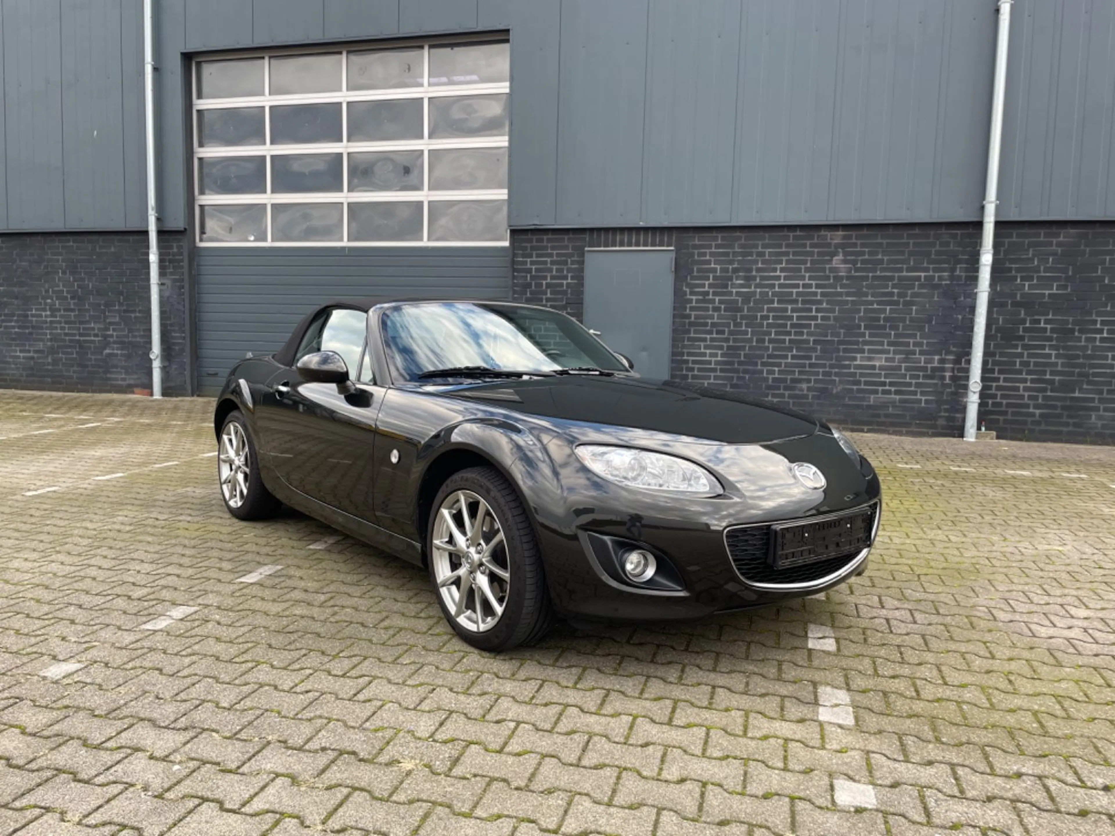 Schwarzer Cabrio-Sportwagen auf einem gepflasterten Parkplatz vor einer modernen, grauen Gebäudefassade geparkt.