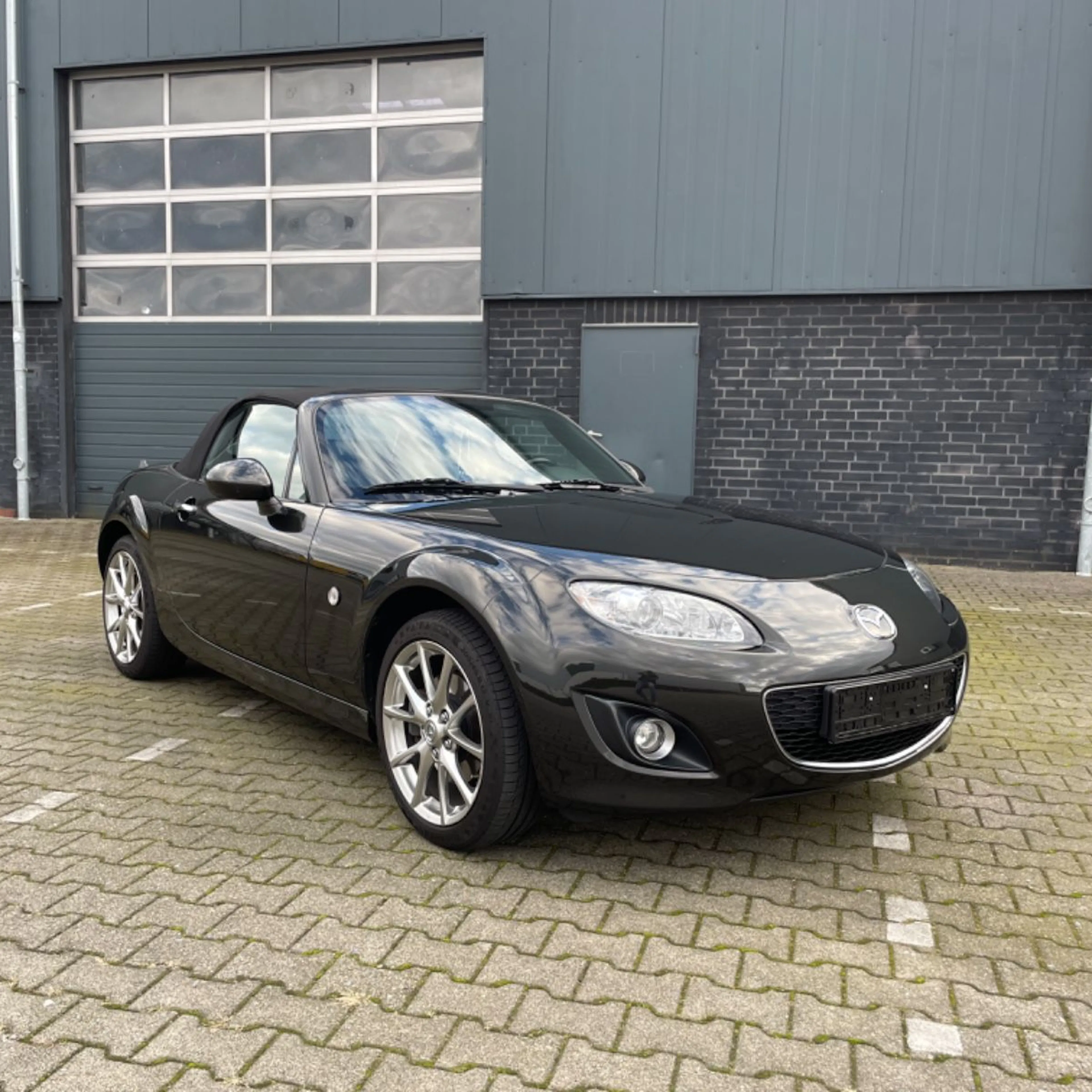 Schwarzer Cabrio-Sportwagen auf einem gepflasterten Parkplatz vor einer modernen, grauen Gebäudefassade geparkt.