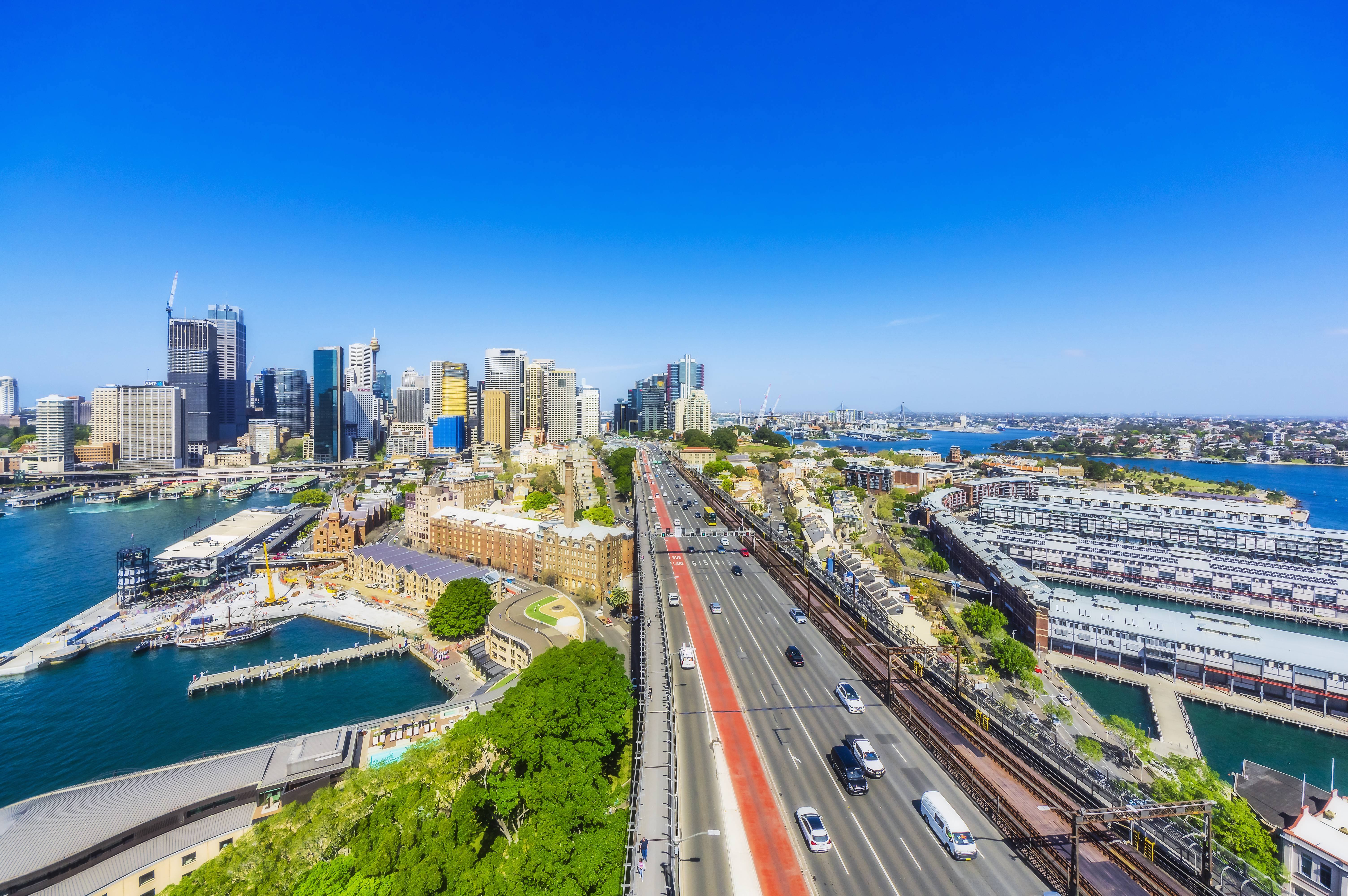 Eine Luftaufnahme von Sydney mit modernen Hochhäusern, einer großen Brücke mit mehreren Fahrspuren und einem angrenzenden Hafenbereich.