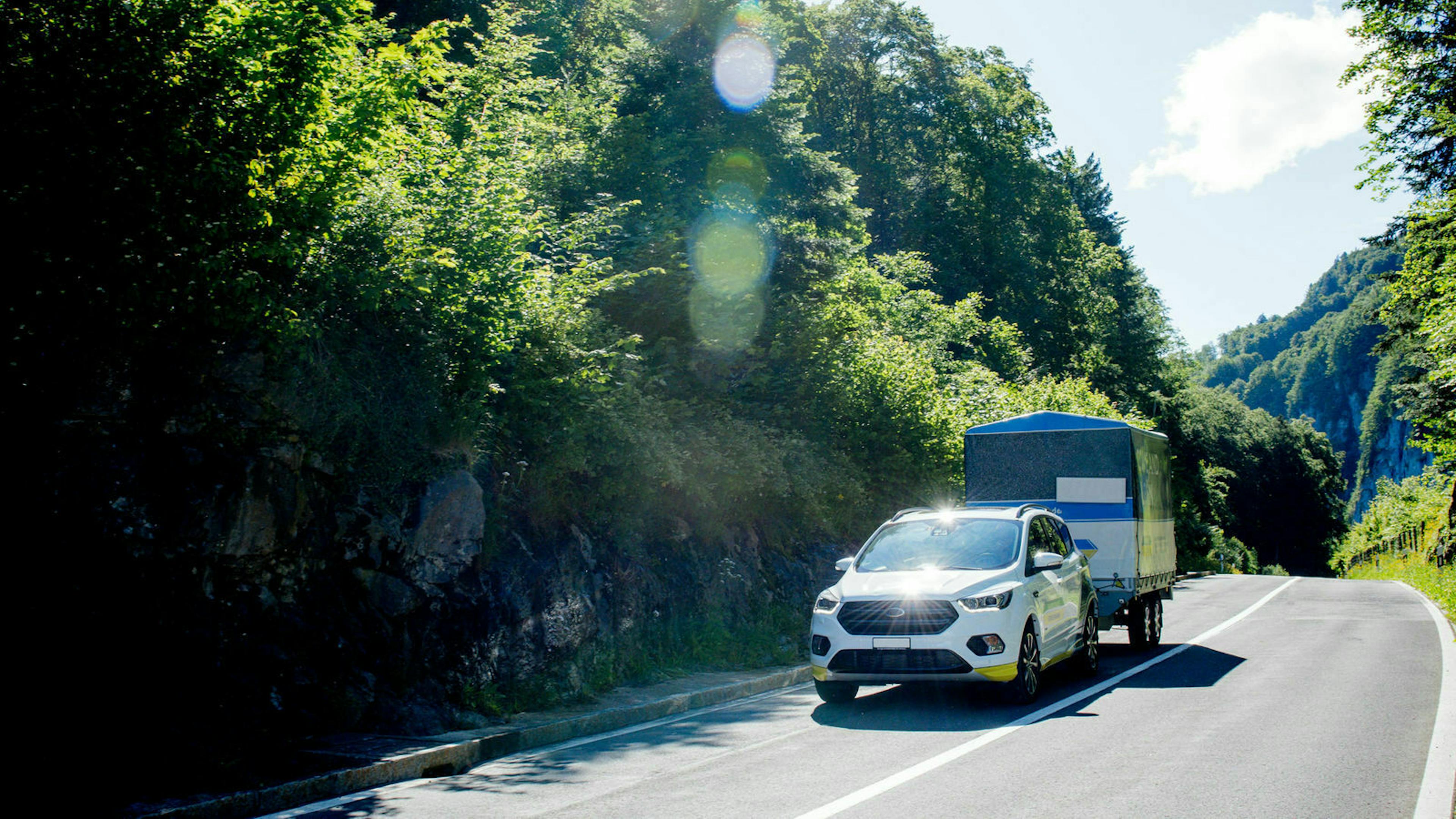 Ein weißer PKW der Marke Ford mit einem blau-weißen Anhänger fährt eine Landstraße entlang