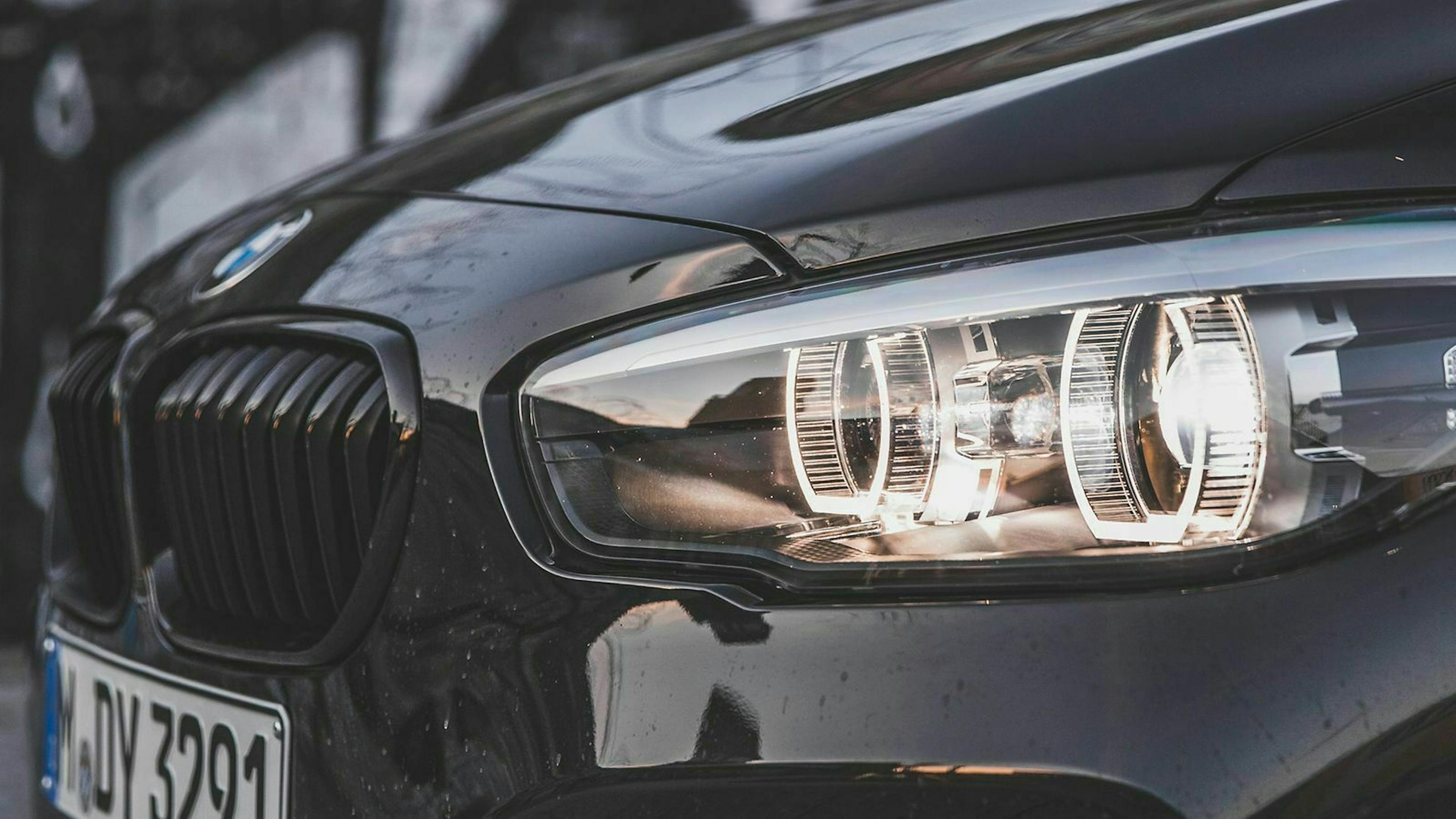 Ein BMW 1er steht mit eingeschaltetem Abblendlicht auf einem Parkplatz. Der Scheinwerfer ist in Nahaufnahme zu sehen. 