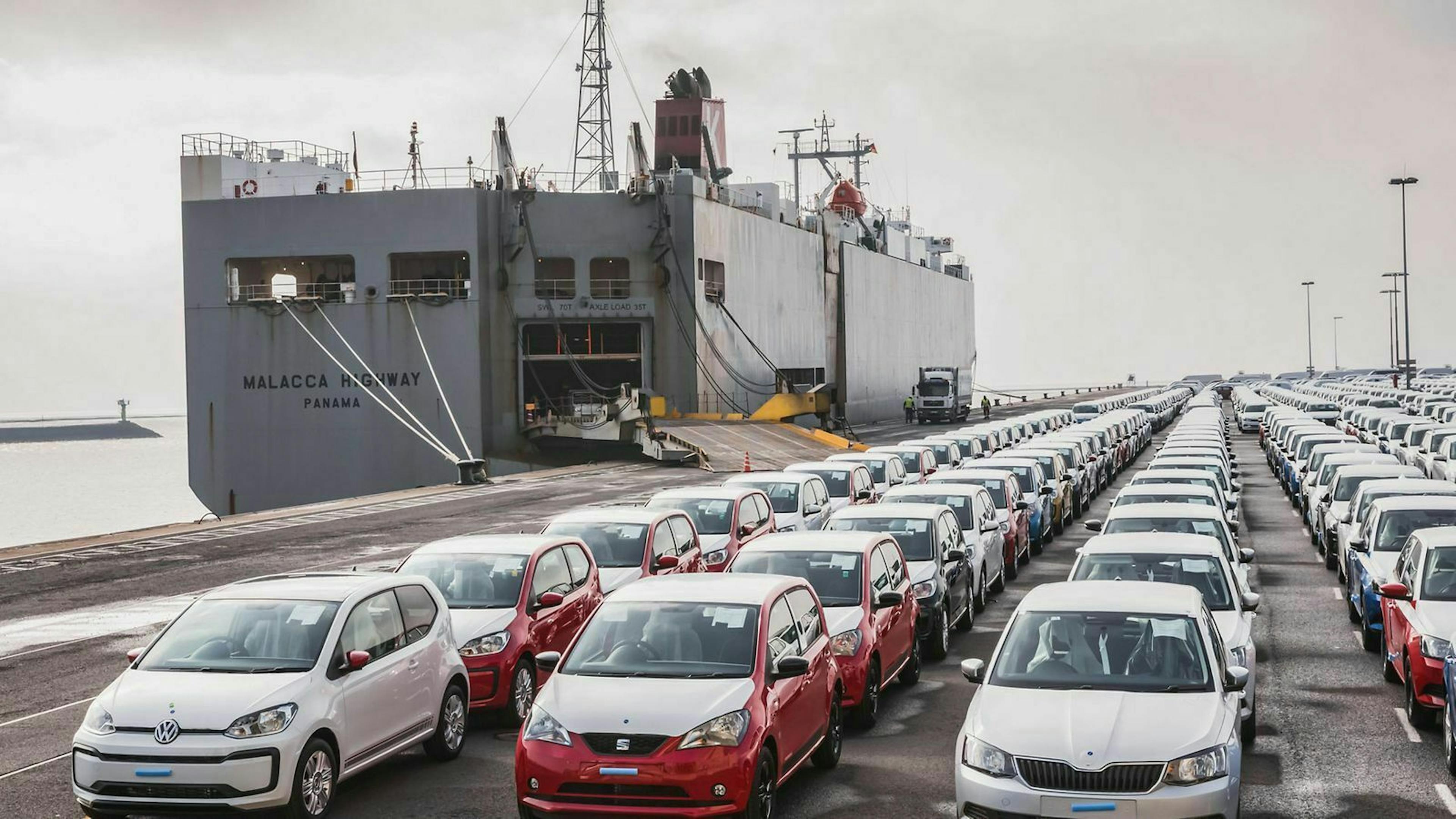 Fahrzeuge stehen am Hafen. Daneben ein Frachtschiff mit offener Ladeluke.