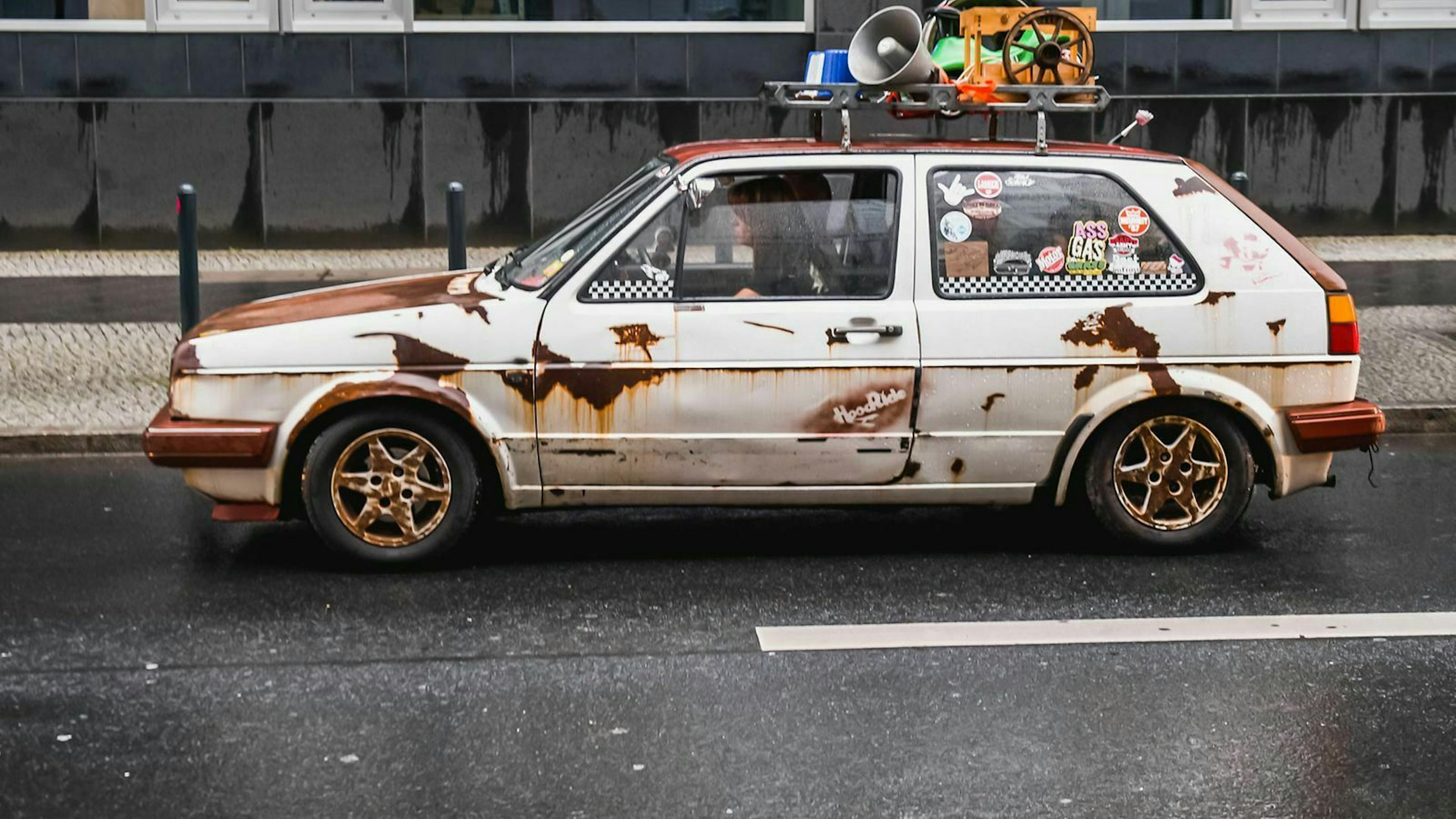 Ein ehemals weißer Golf II mit bewusst gesetzten Rostflächen fährt durchs Bild.