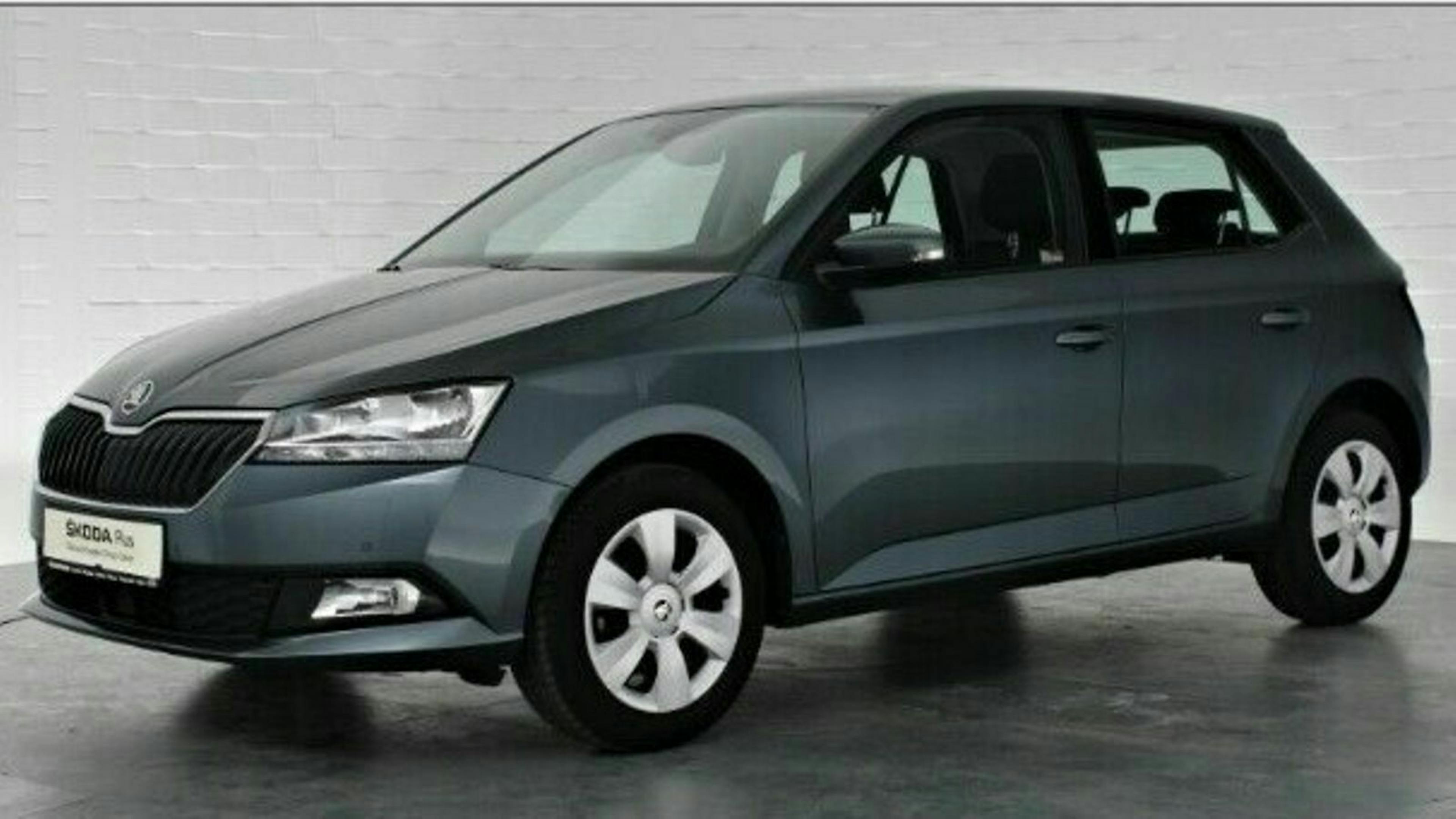 Grauer Škoda Fabia steht in einem hellen Raum vor einer weißen Ziegelwand, leicht seitliche Ansicht, Vorderteil gut sichtbar.