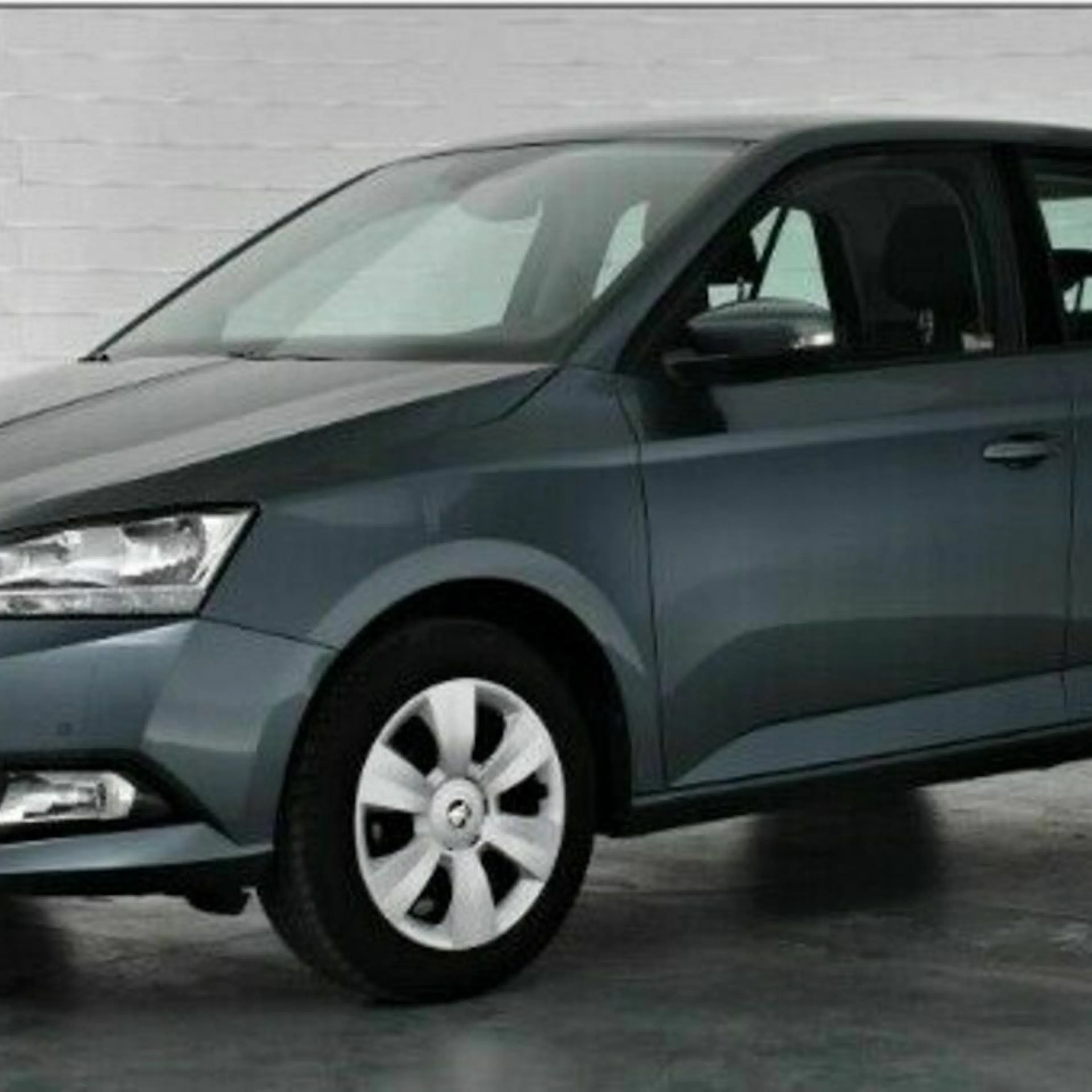 Grauer Škoda Fabia steht in einem hellen Raum vor einer weißen Ziegelwand, leicht seitliche Ansicht, Vorderteil gut sichtbar.