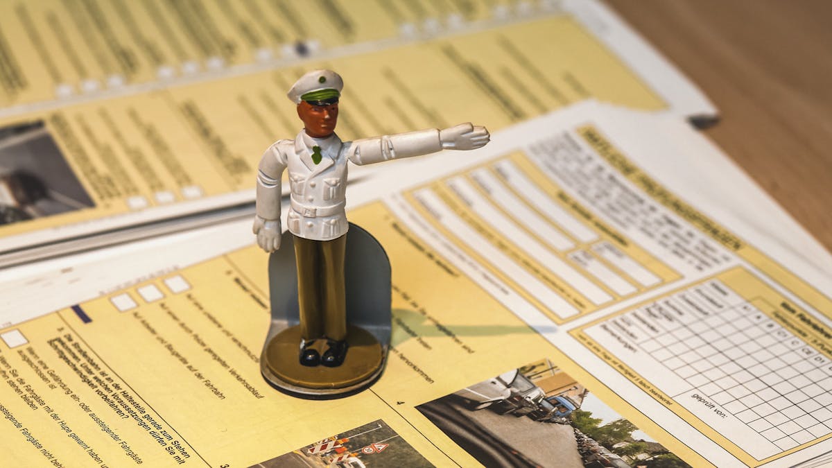 Die Modellfigur eines Streifenpolizisten steht auf einem Fahrschul-Prüfungsbogen und weist eine Richtung