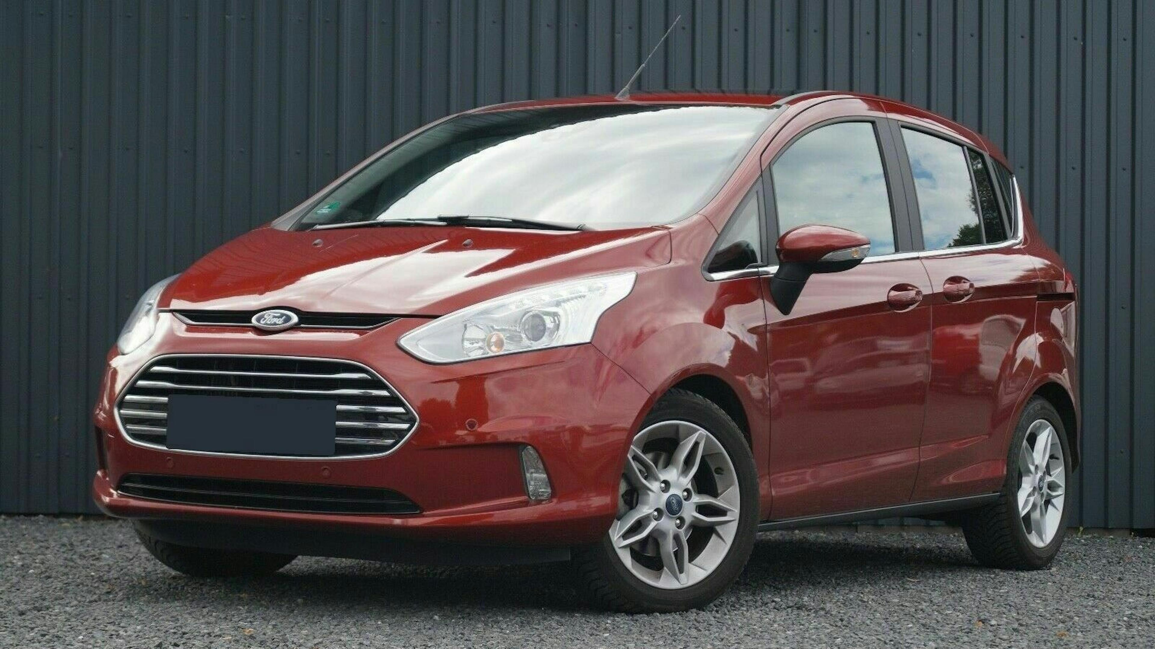Roter Ford-B-Max-Kleinwagen auf Schotter vor einer schwarzen, geriffelten Wand geparkt. Frontansicht bei Tageslicht.