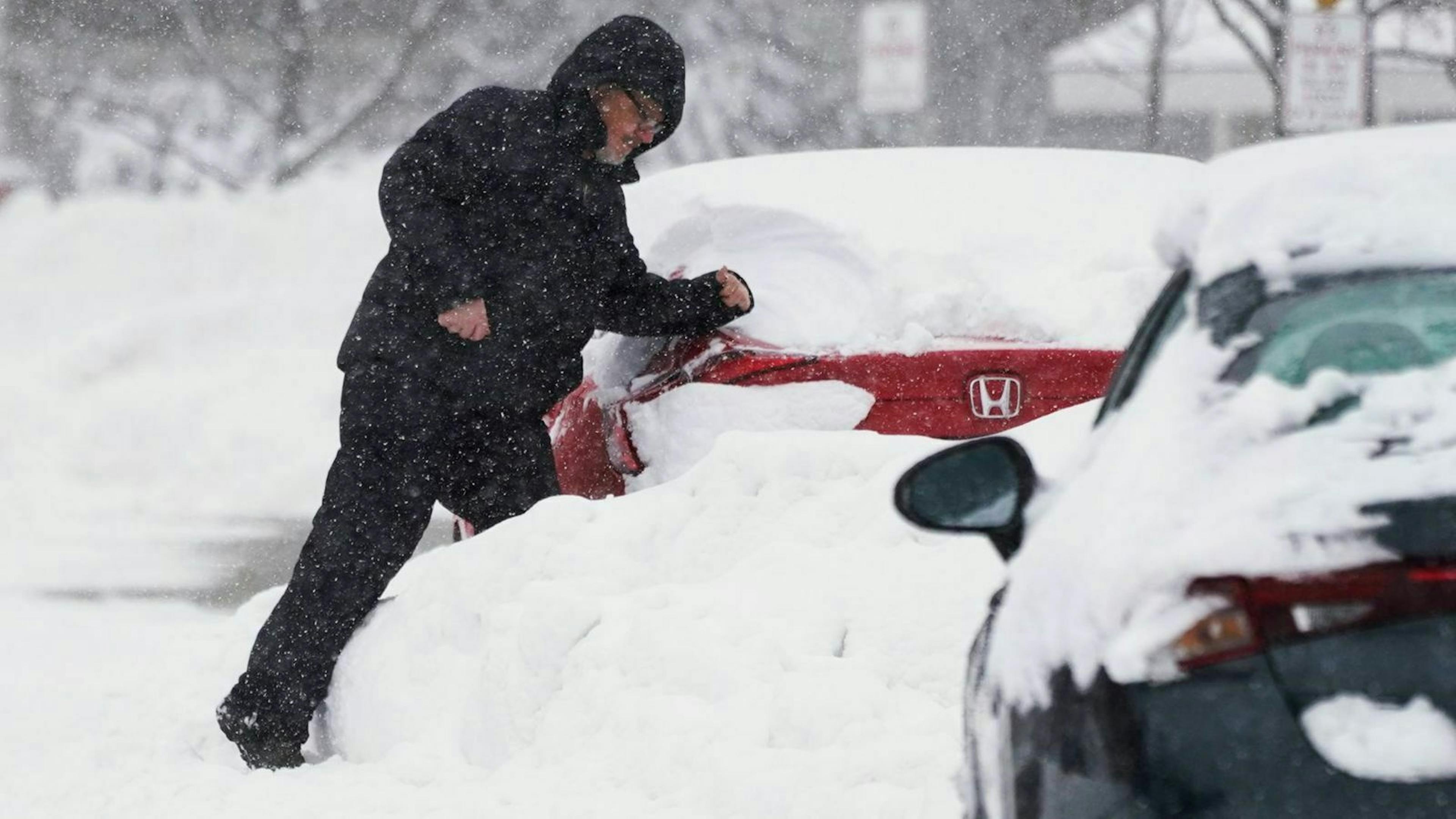 Zu sehen ist ein Mann, der Schnee von seinem Auto mit dem Arm wegschiebt