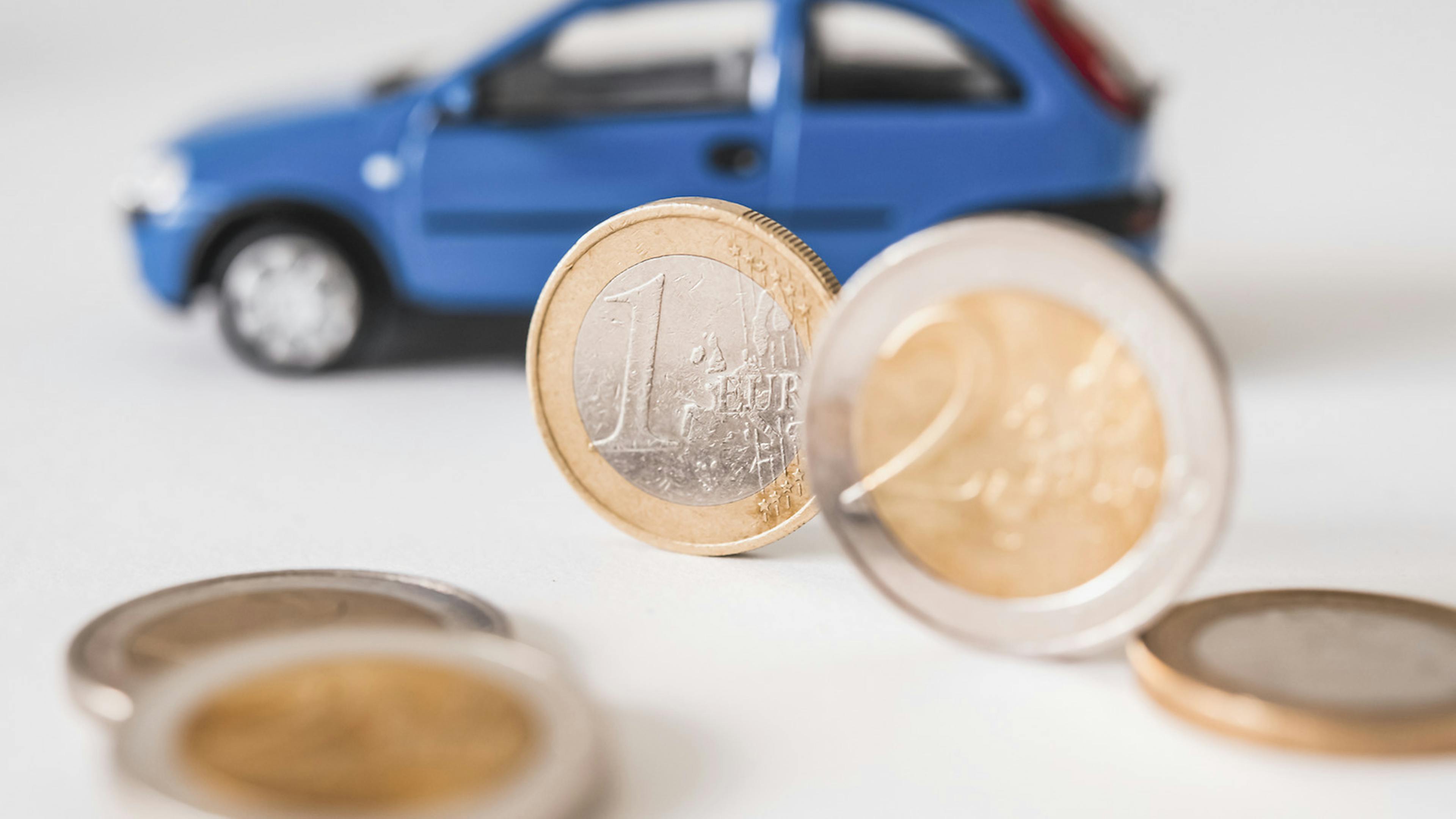 Eine 1-Euro und eine 2-Euro-Münze stehen aufrecht vor einem blauen Modellauto eines Kleinwagens