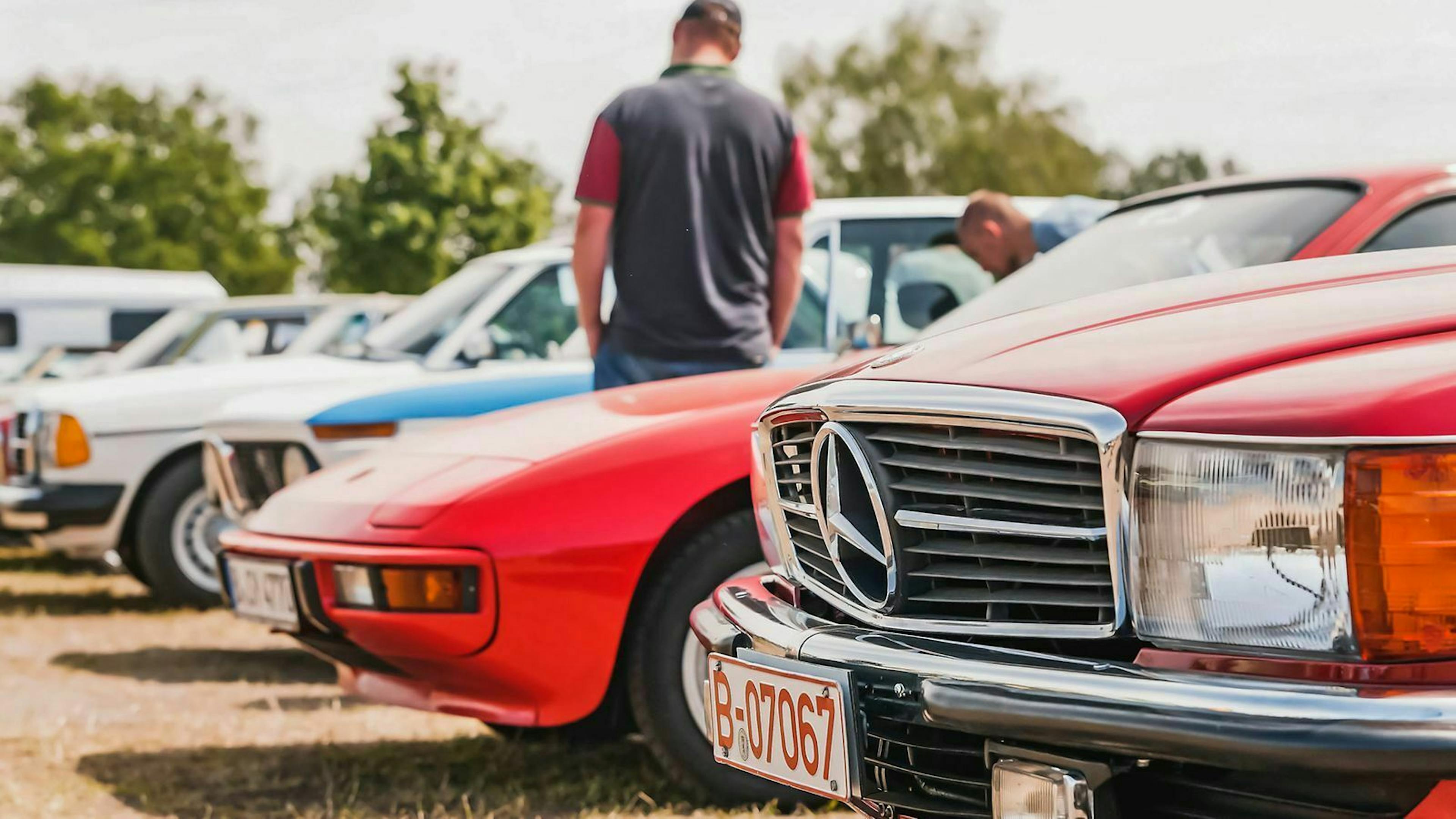 Im Vordergrund steht ein mercedes Roadster, im Hintergrund weitere Youngtimer-Sportwagen, die von einem Mann begutachtet werden.