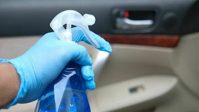 Detailaufnahme einer Hand, die eine Sprühflasche mit Reinigungsmittel hält, im Hintergrund ist der Innenraum eines PKW