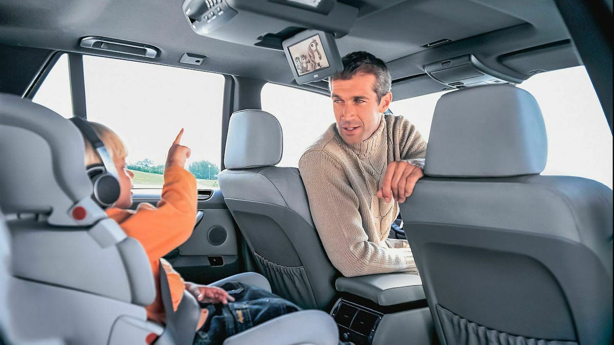 Ein Kind im Kindersitz auf der Autorückbank zeigt auf den Monitor am Fahrzeughimmel während sich der Fahrer zum Kind umschaut.