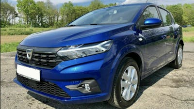 Ein blauer Dacia Sandero steht auf einem Parkplatz vor einer Baumreihe.