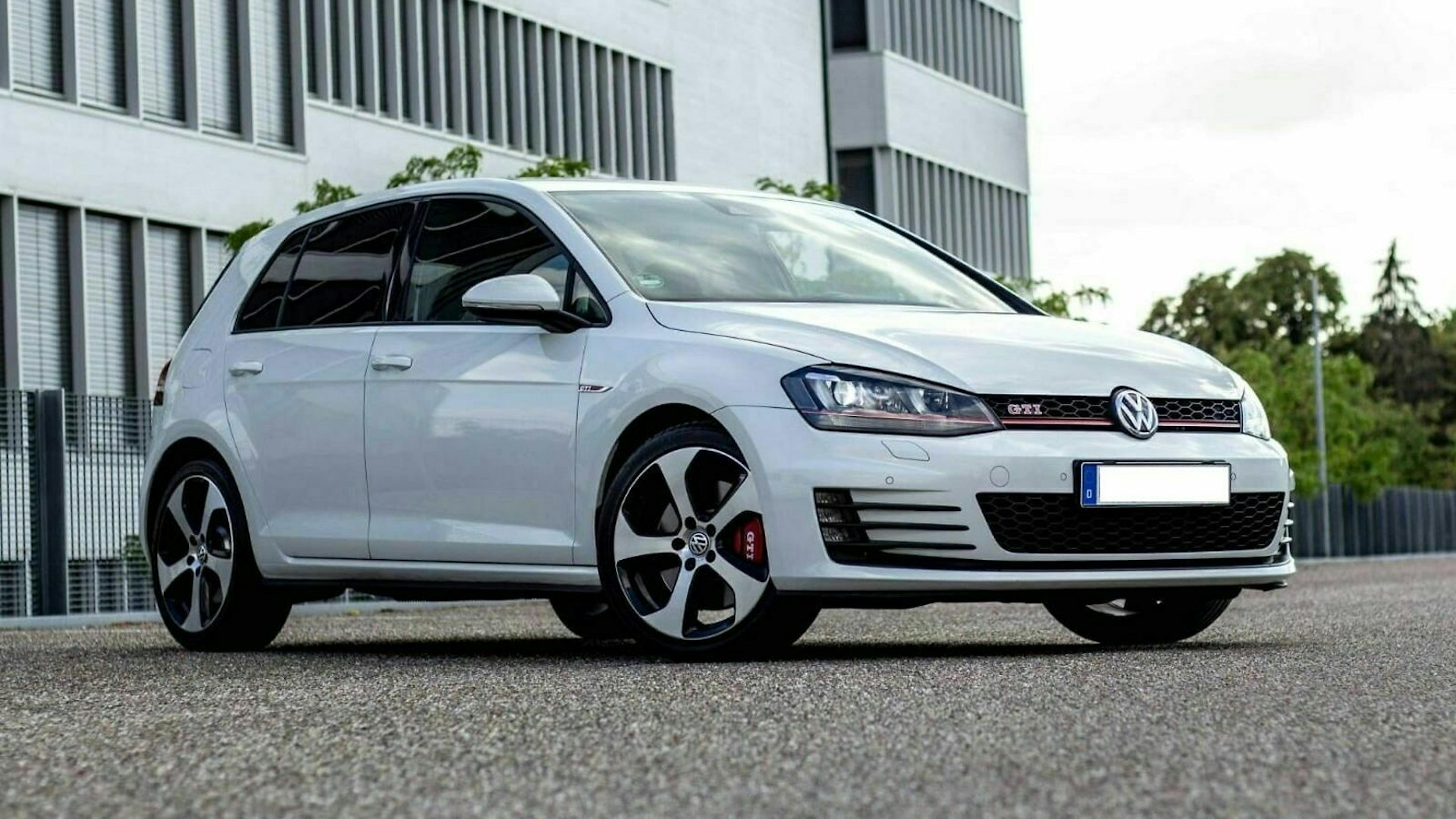 Weißer Volkswagen Golf GTI parkt vor einem modernen Gebäude; sportliches Design mit markanten Felgen und getönten Scheiben.
