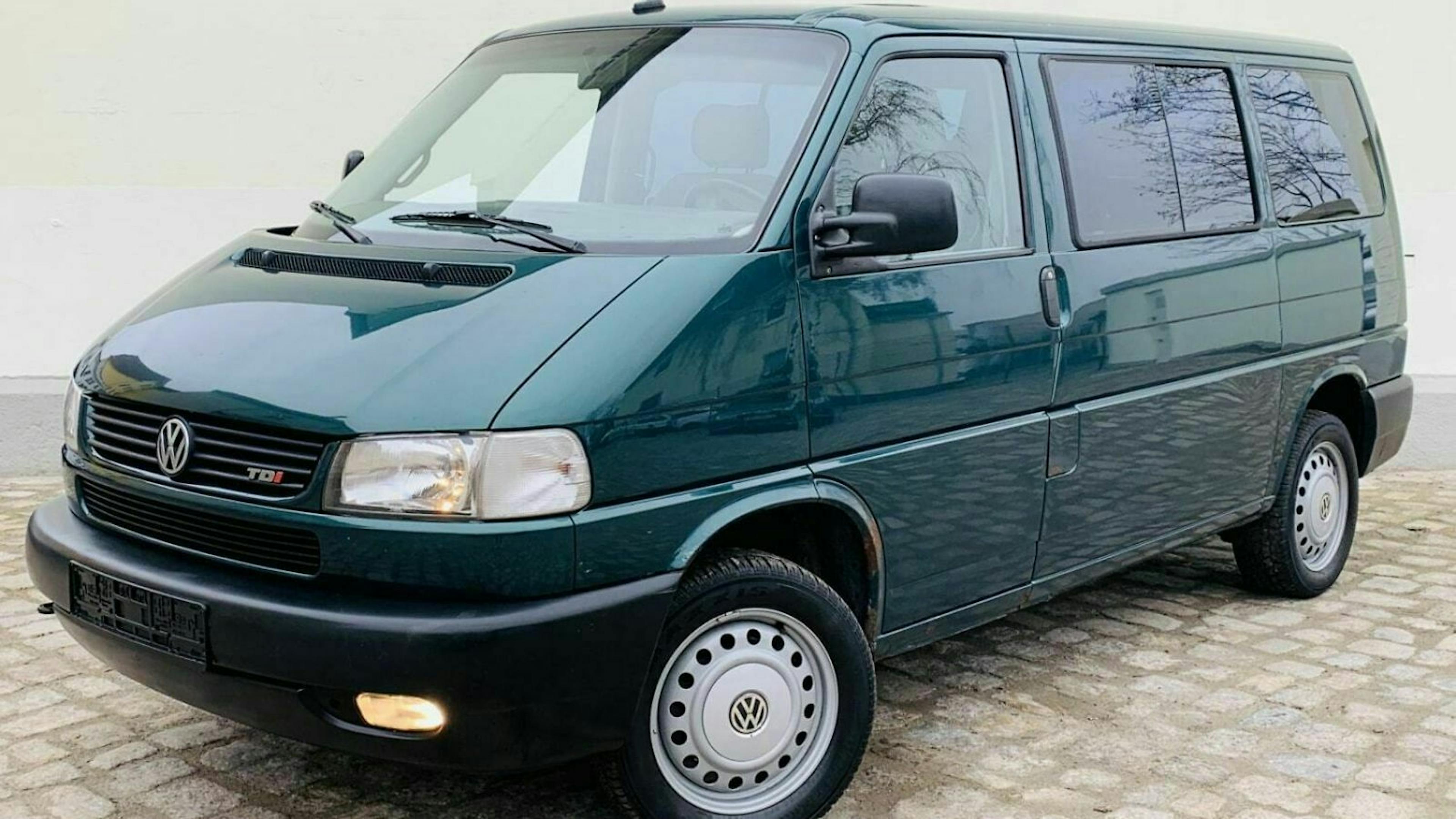 Dunkelgrüner VW T4 Transporter, seitlich fotografiert, auf einer gepflasterten Fläche stehend. Vorderlichter eingeschaltet, klare Fenster.