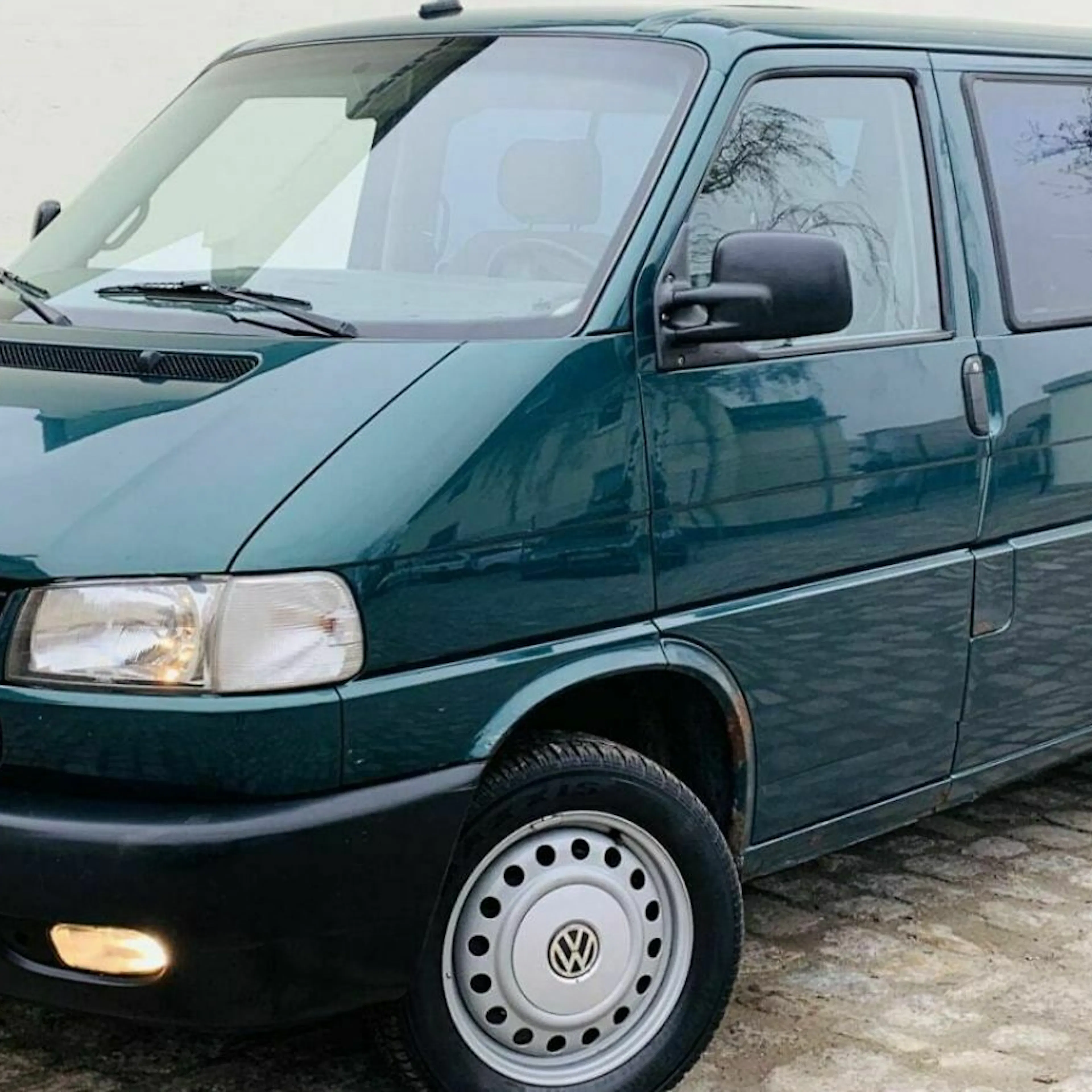 Dunkelgrüner VW T4 Transporter, seitlich fotografiert, auf einer gepflasterten Fläche stehend. Vorderlichter eingeschaltet, klare Fenster.