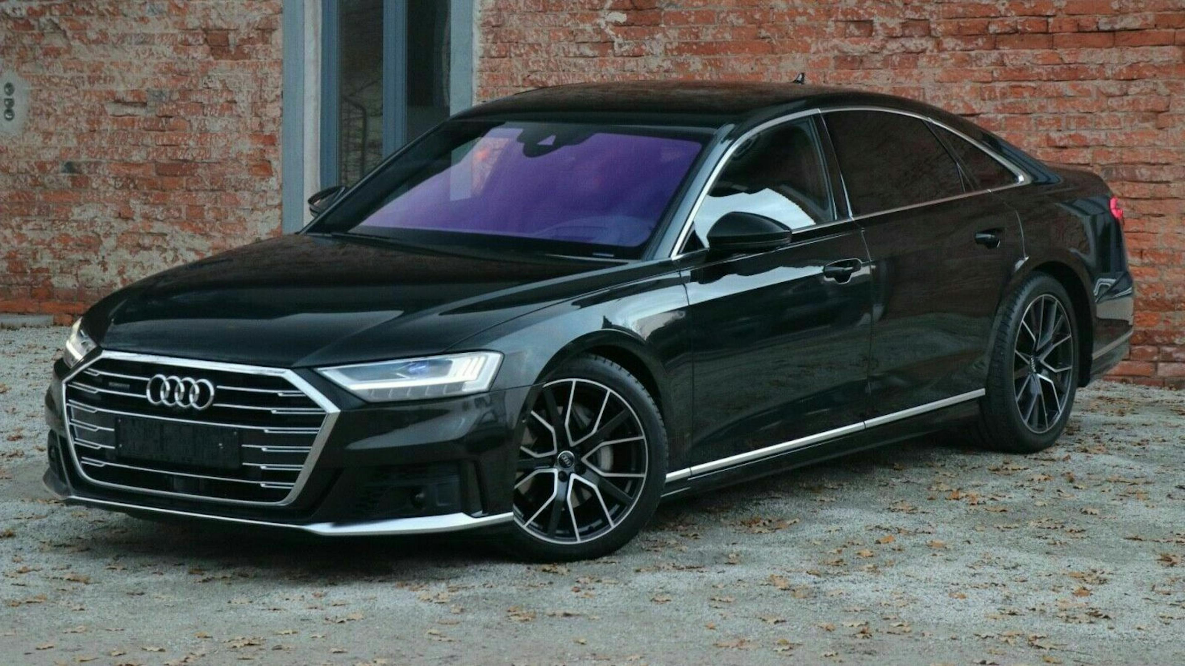 Schwarzer Audi A8 vor einer roten Backsteinwand geparkt. Moderner Stil mit markanten Scheinwerfern und glänzenden Leichtmetallrädern.