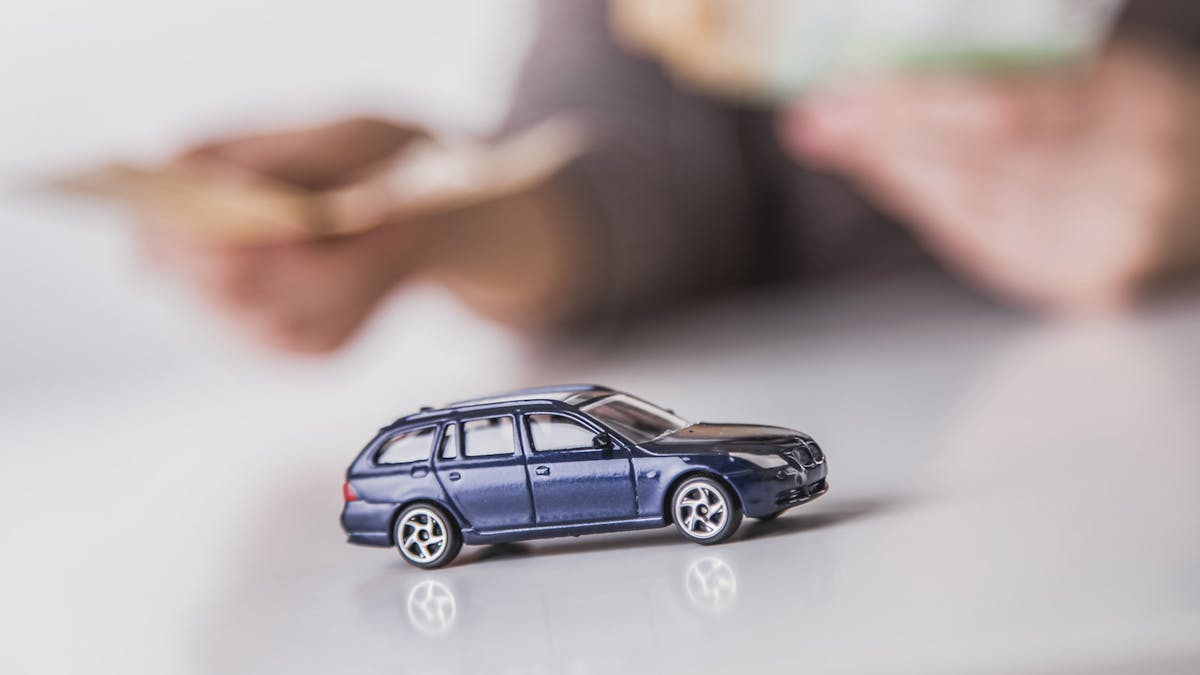 Ein blaues Auto steht auf einem weißen Tisch. Unscharf im Hintergrund zählt jemand Geldscheine