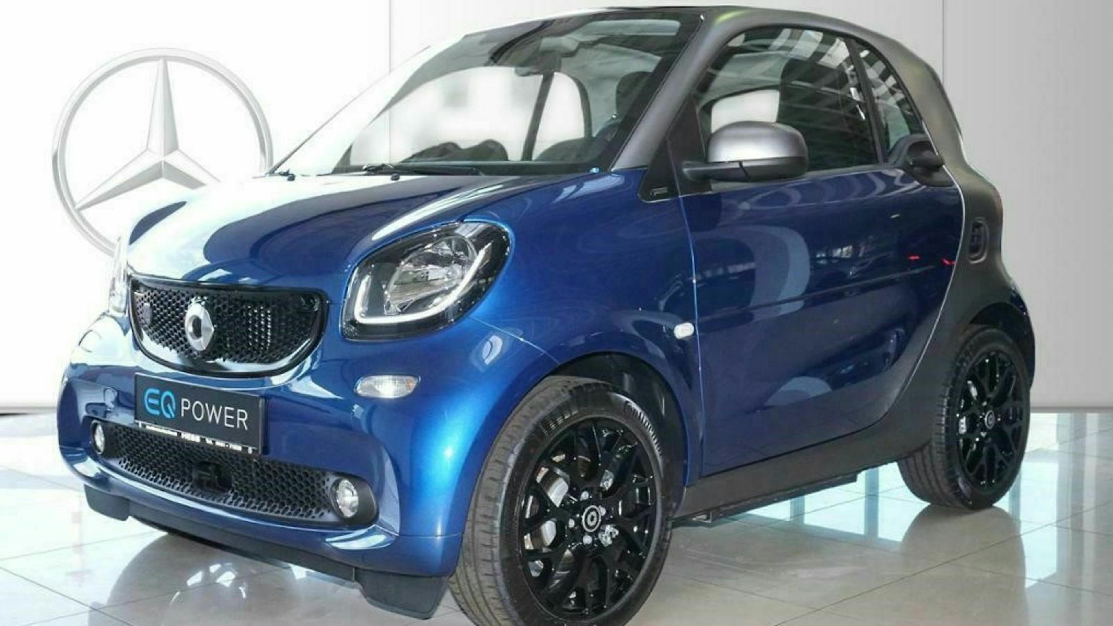 Blauer Smart EQ Fortwo mit schwarzem Dach in einem Showroom, sichtbar durch Mercedes-Logo im Hintergrund. Fahrzeug steht auf glänzendem Boden.
