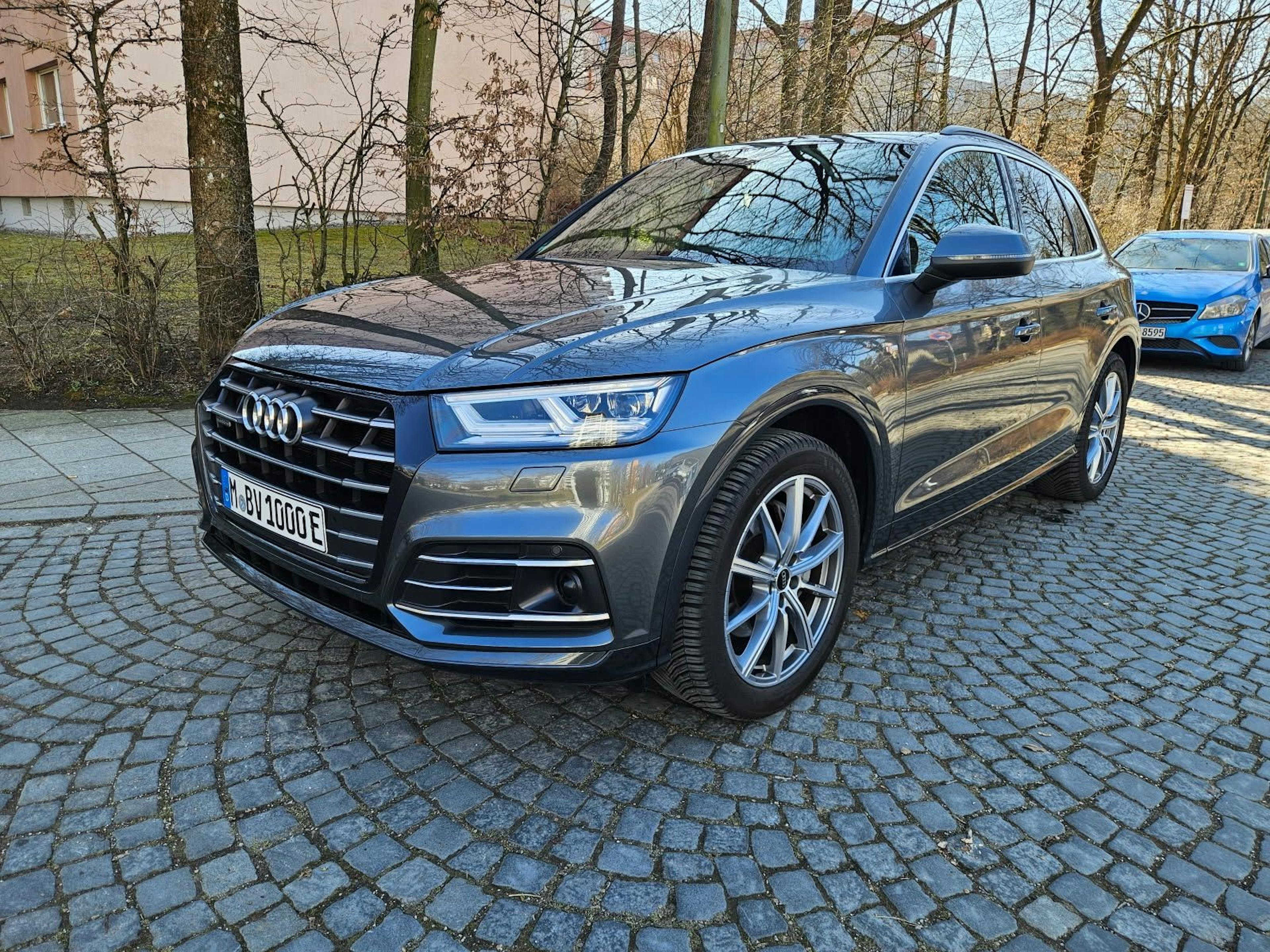Grauer Audi Q5 Plug-in-Hybrid mit Münchner Kennzeichen, geparkt auf einem Kopfsteinpflasterweg in einer baumbestandenen Umgebung.