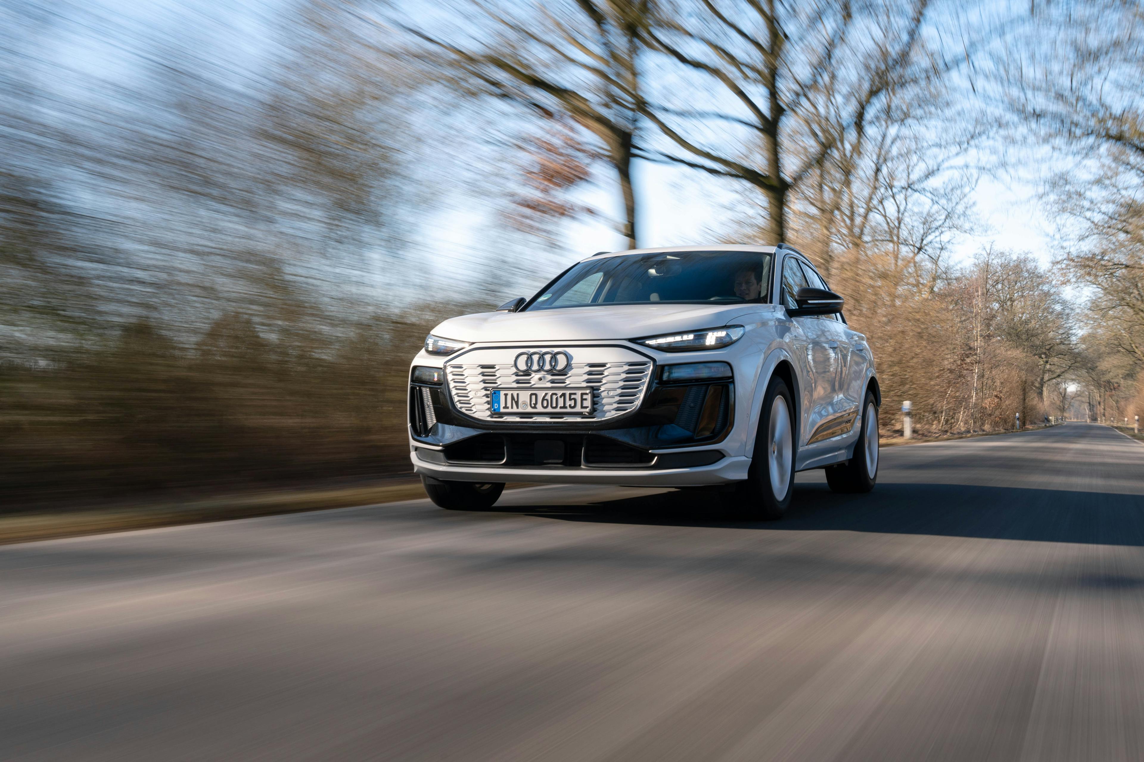 Ein weißer Audi Q6 e-tron quattro fährt auf einer Landstraße vor laublosen Bäumen.