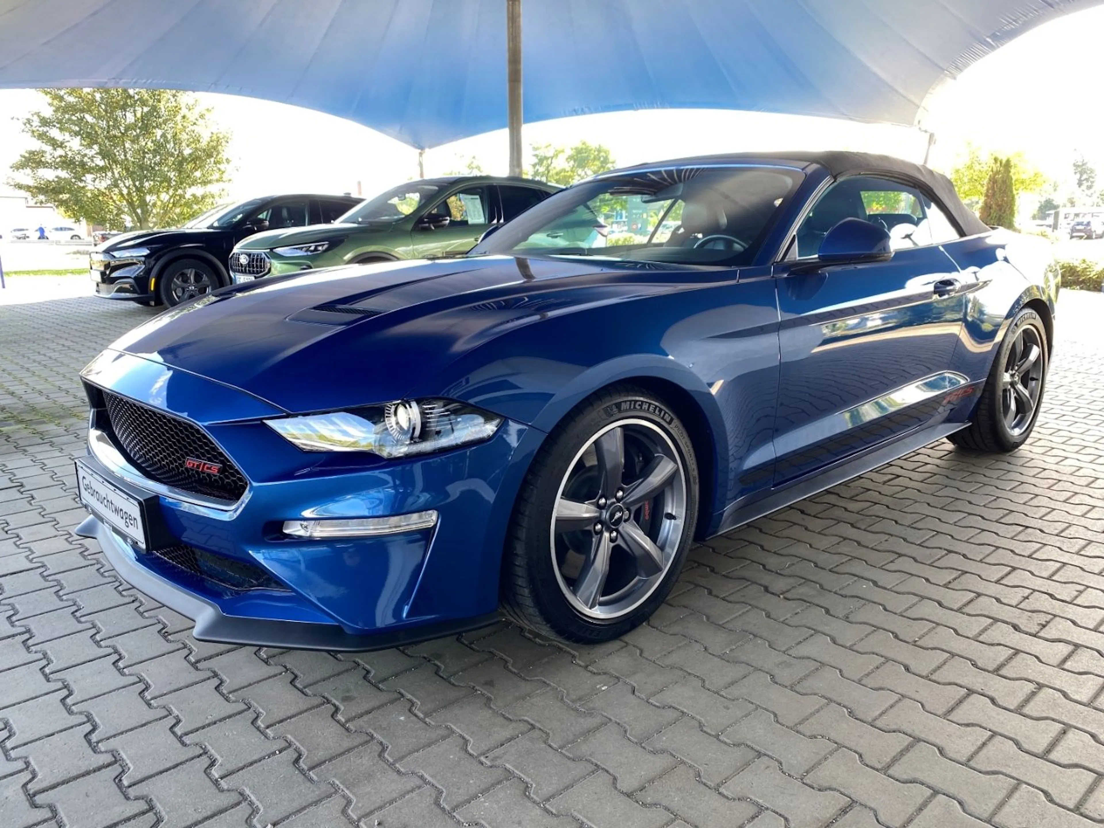 Blauer Sportwagen mit offenem Verdeck unter einem großen Pavillon geparkt. Weitere Autos im Hintergrund.