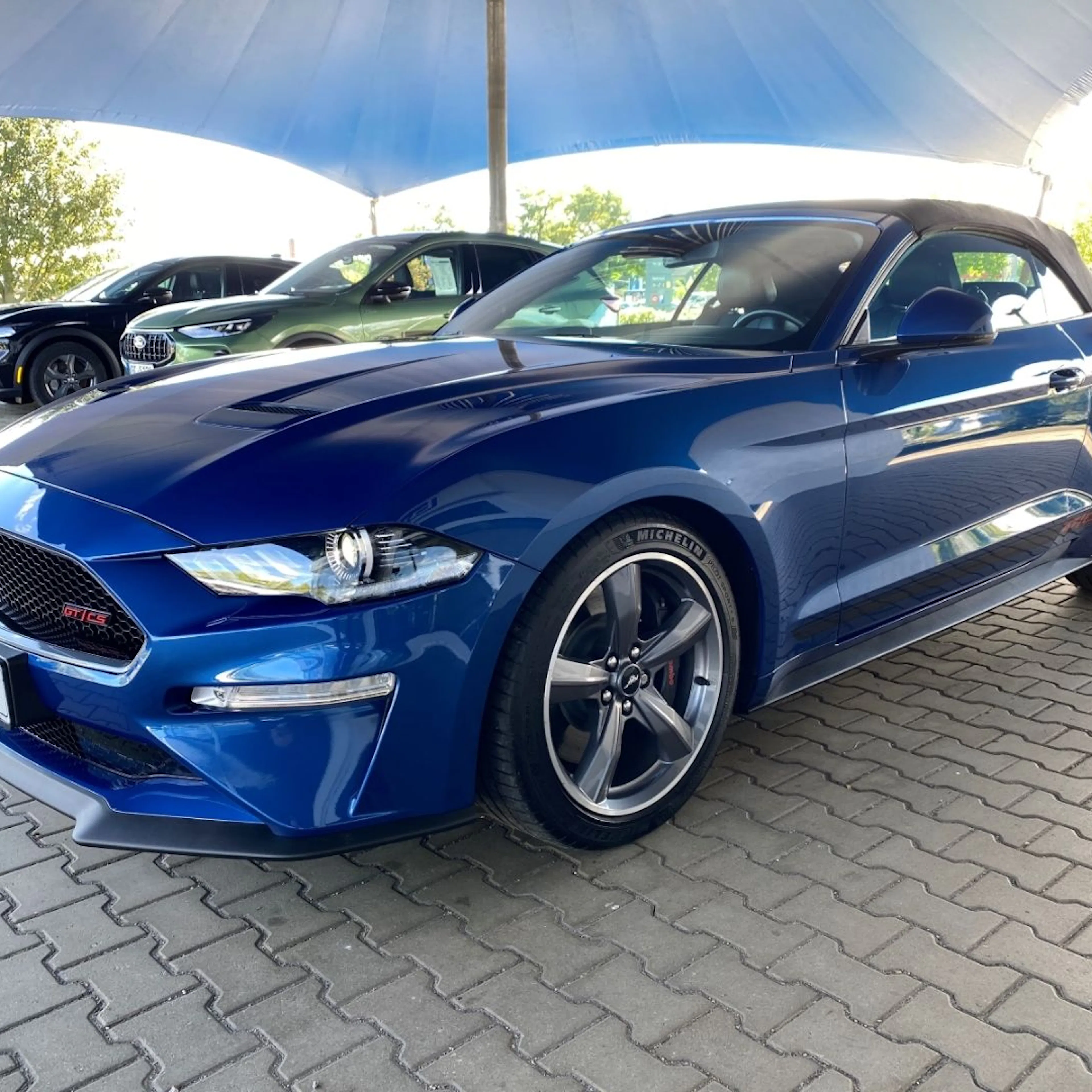 Blauer Sportwagen mit offenem Verdeck unter einem großen Pavillon geparkt. Weitere Autos im Hintergrund.