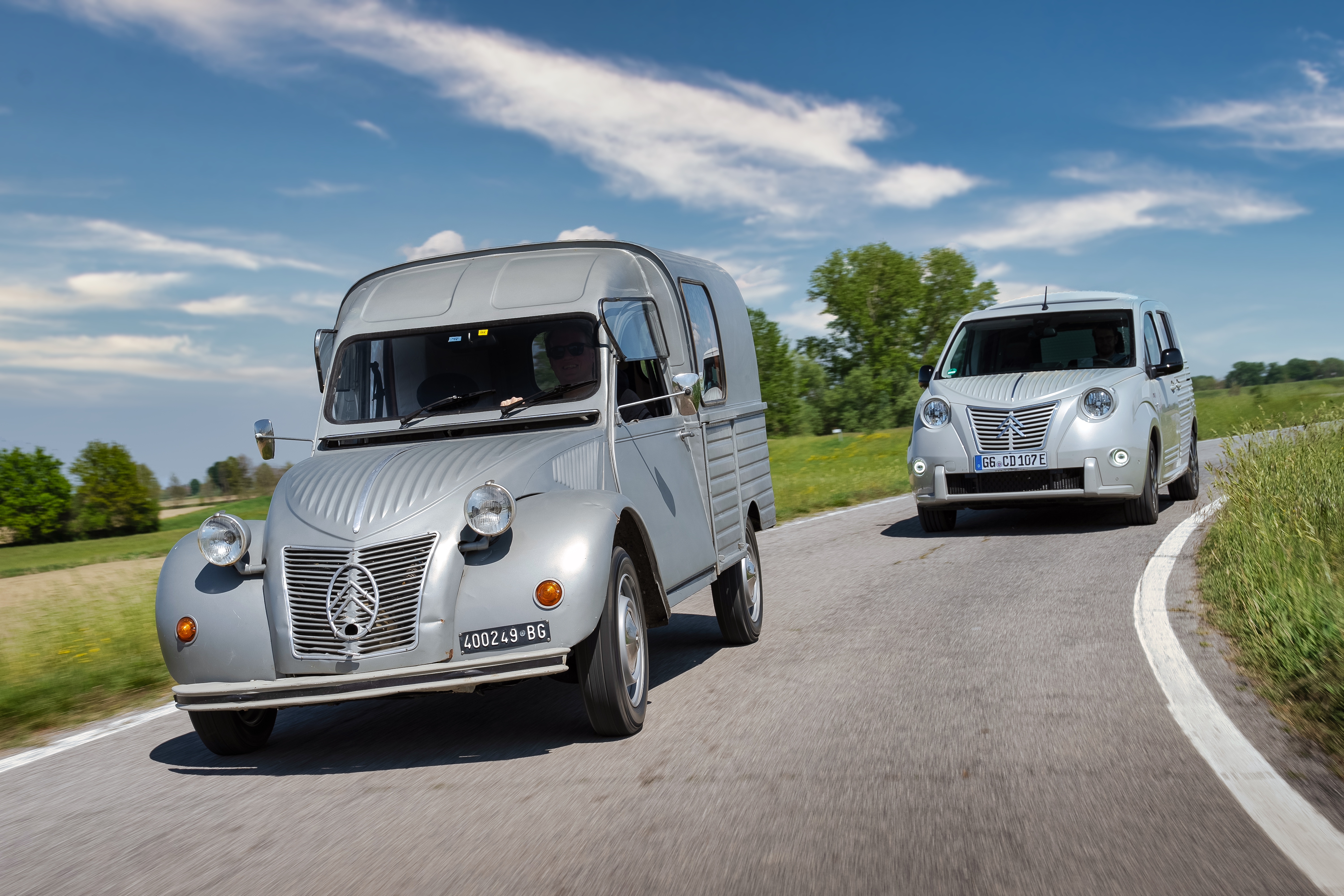 Graue Citroën 2CV Kastenente fährt auf einer Landstraße, dahinter ein silberner Berlingo mit Doppelwinkel-Kühlergrill.
