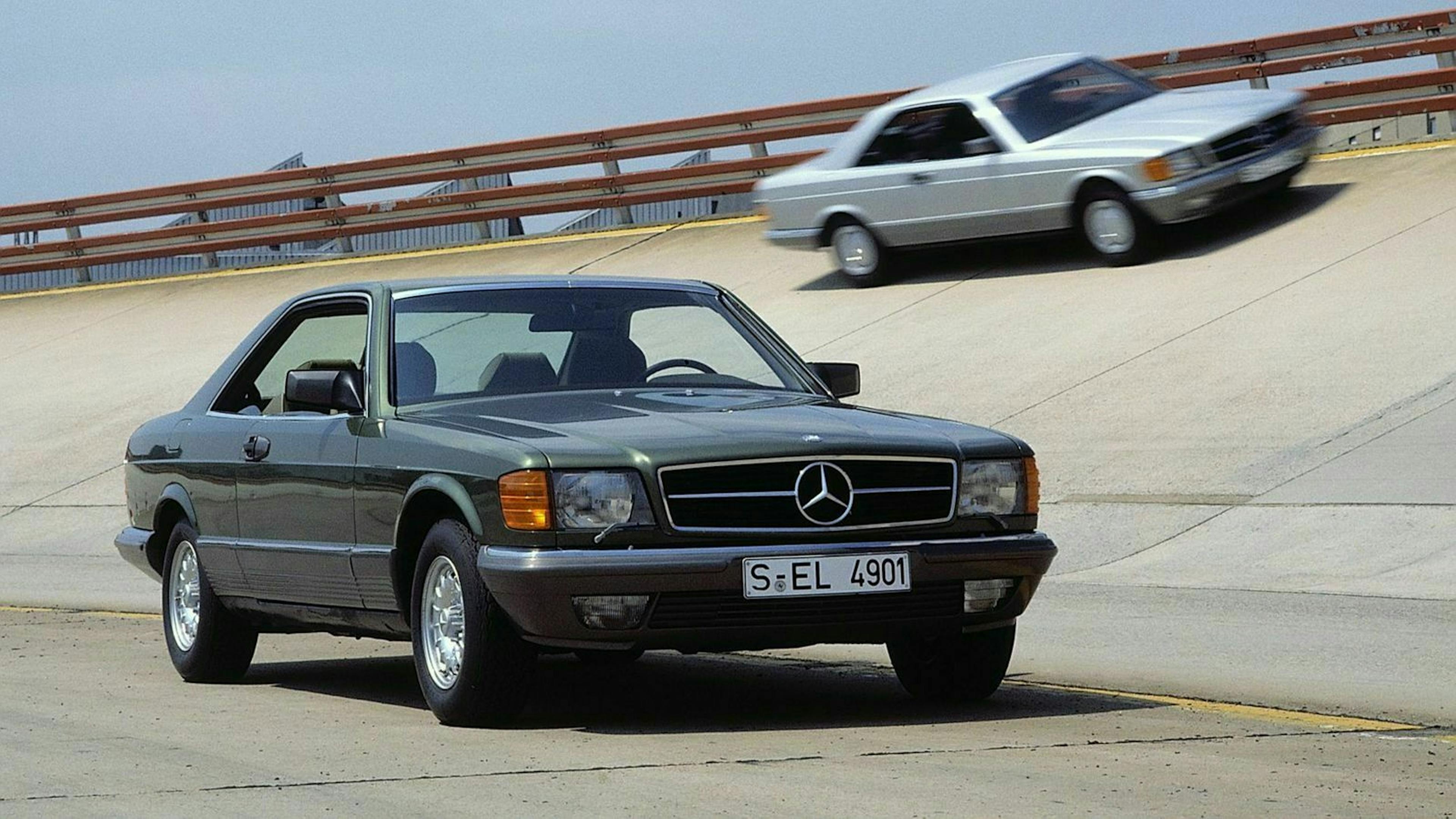 Zwei Mercedes-Oldtimer auf einer Straße; im Vordergrund ein grüner Wagen, im Hintergrund ein silberner Wagen auf einer schiefen Ebene.